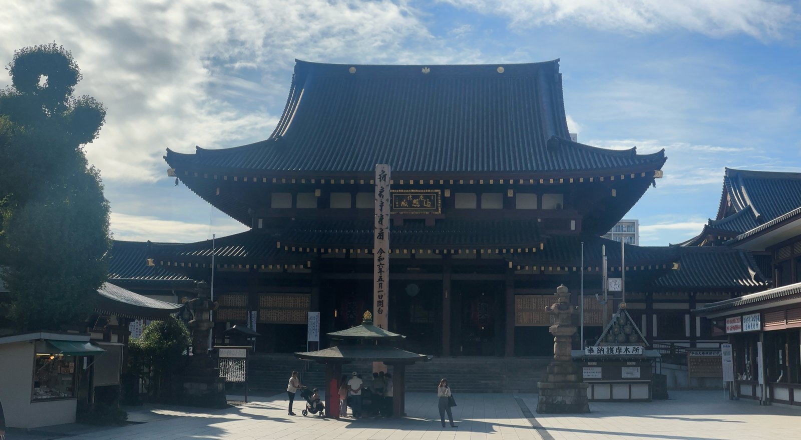 川崎大師の大本堂。大きな瓦屋根の木造の堂で、正面の石畳の広場に参拝者が数人いる。手前に屋根付きの香炉と石灯籠