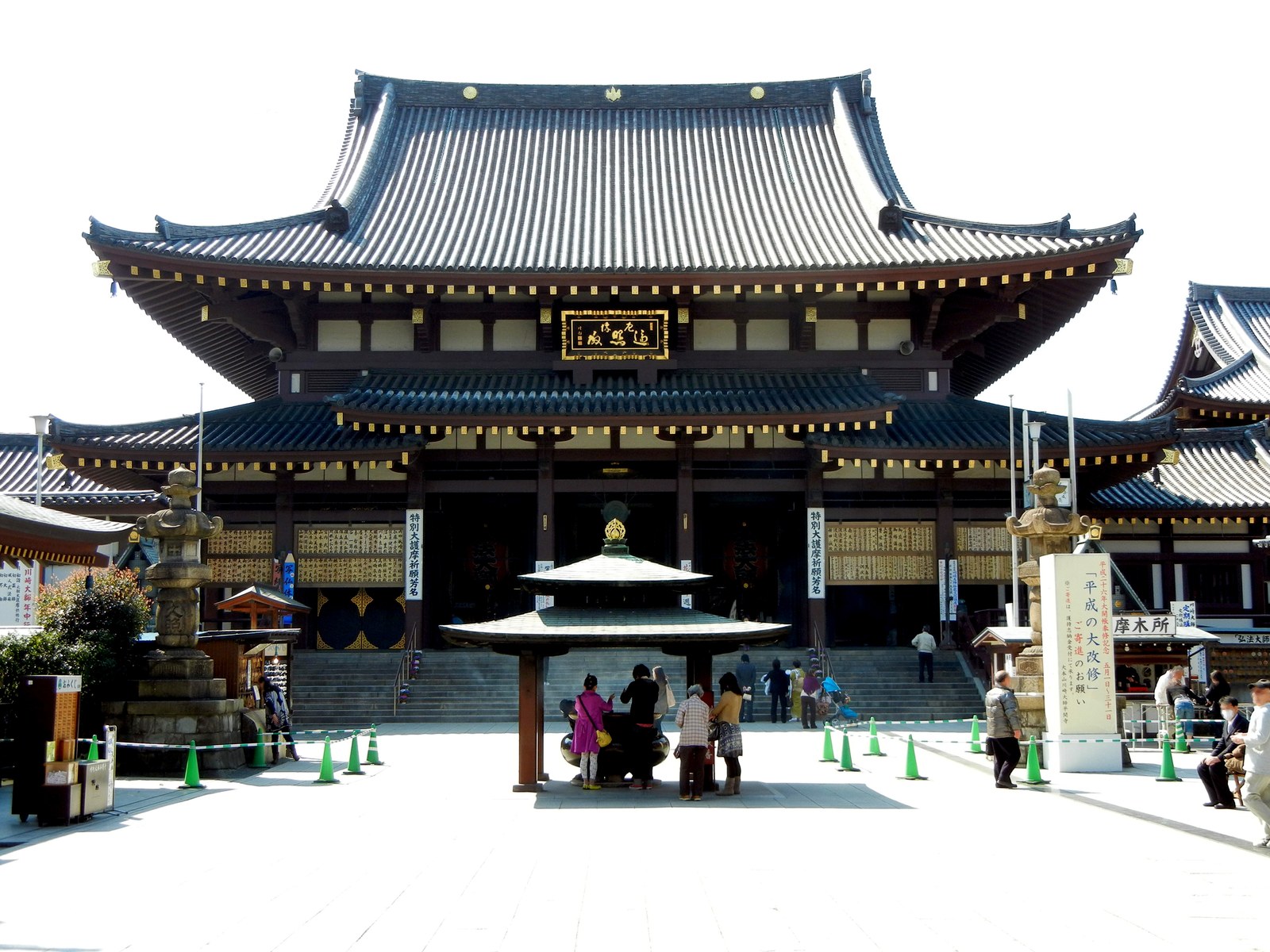 川崎大師の大本堂。二重の瓦屋根の大きな本堂の前に香炉があり、参拝の人が数人立っている