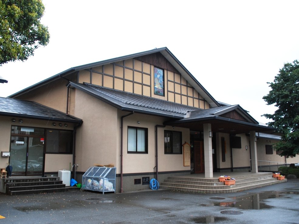 雨にぬれた駐車場の奥に、瓦屋根の2階建て風の大きな建物が立つ。入口に石段と柱の庇があり、軒先に花の鉢が並んでいる