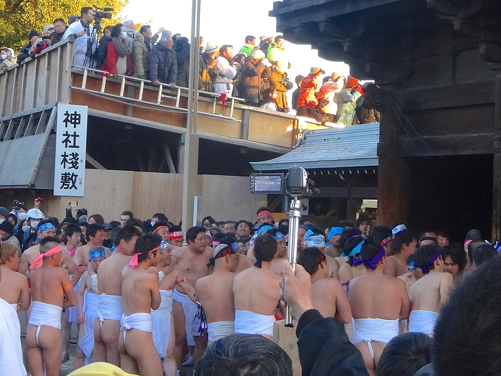 国府宮はだか祭。白いさらしを締め、色のはちまきをした裸男たちが門の前に集まり、左上の「神社桟敷」の看板の下に見物の人が並ぶ