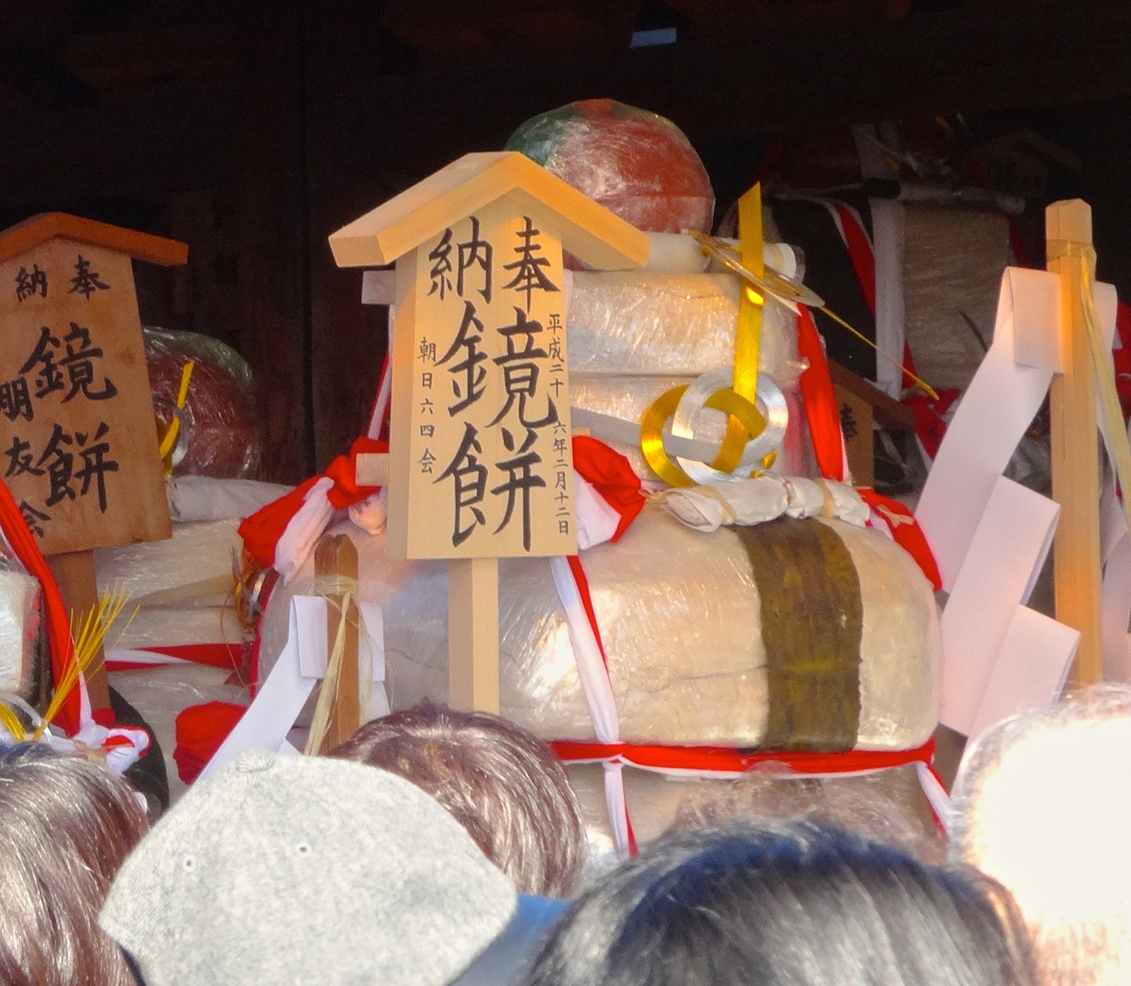 奉納された鏡餅。透明な包みの餅を重ね、上に橙を載せ、紅白の布と金銀の水引で飾り、「奉納 鏡餅」と書いた木の札が立つ