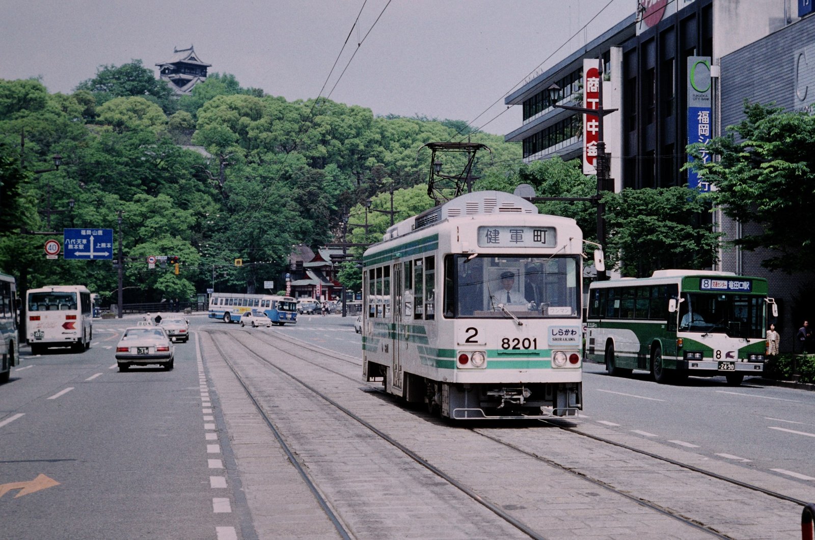 緑の帯の白い路面電車8201が「健軍町」の表示を出して大通りを走り、奥の緑の丘の上に熊本城が見える