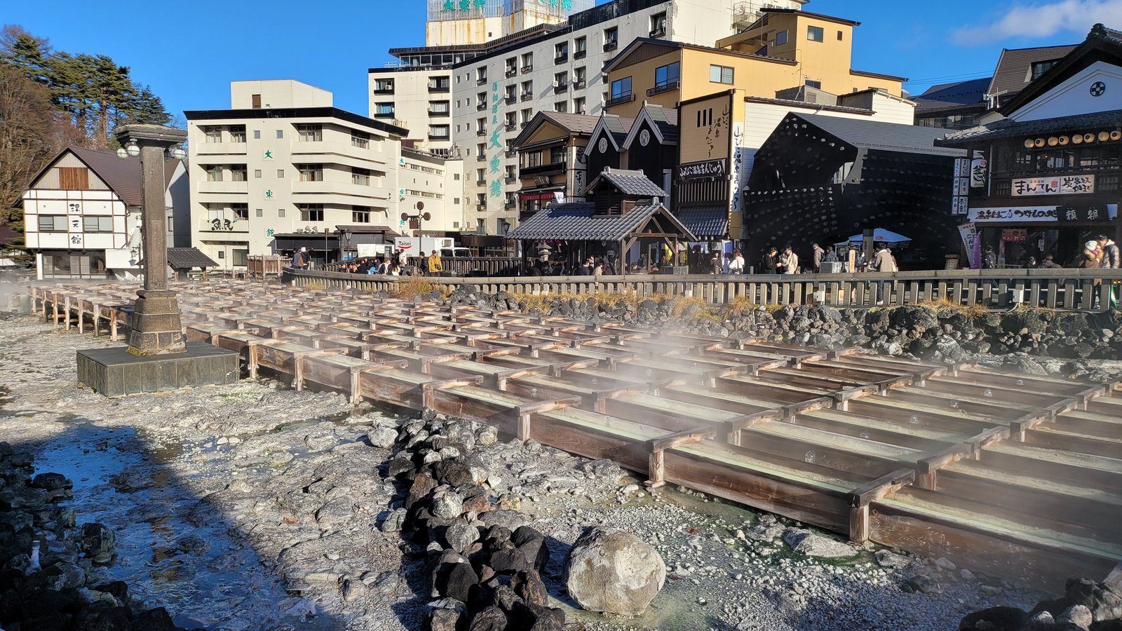 草津温泉の湯畑。木の湯樋が並び、湯けむりが上がる。奥にホテルや土産物店の建物が並び、柵の外に見物の人が立っている。