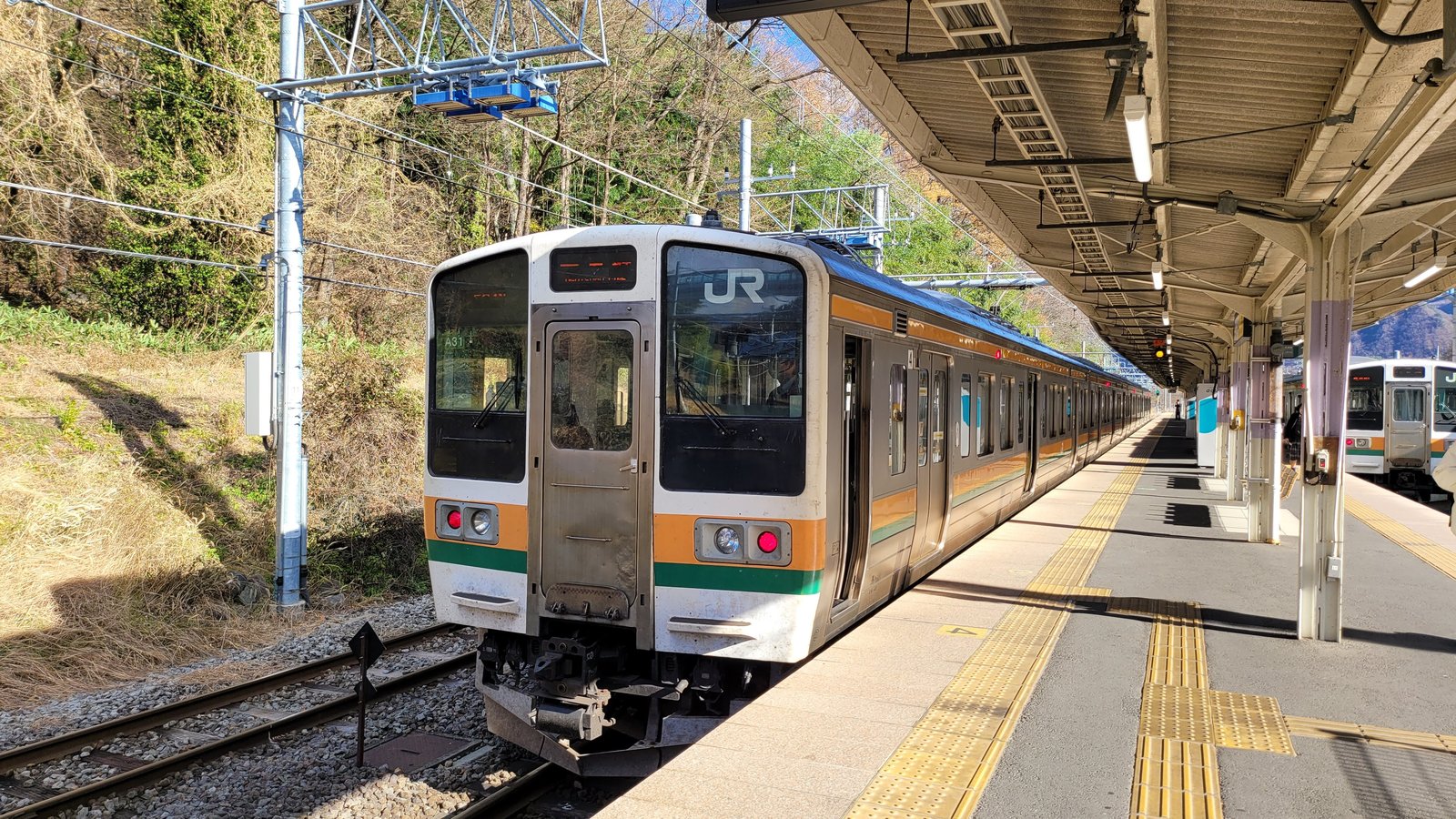 長野原草津口駅のホームに停まる、白地にオレンジと緑の帯の JR 普通列車。右側に屋根付きのホームと、隣のホームにもう1編成が見える
