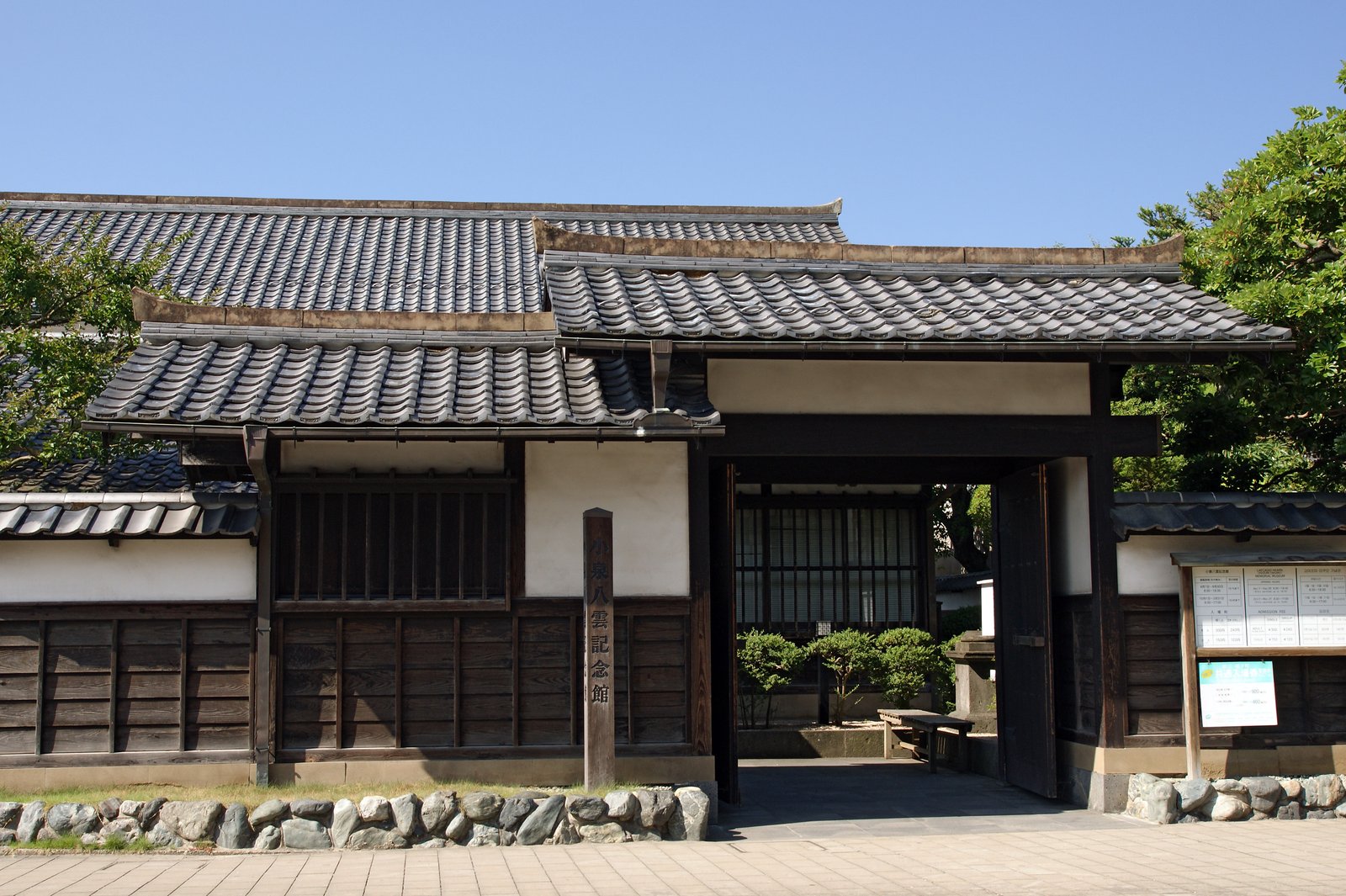 小泉八雲記念館の門。黒い板塀と白い漆喰の壁に瓦屋根の門が開き、門の前に「小泉八雲記念館」と彫った木の標柱が立つ