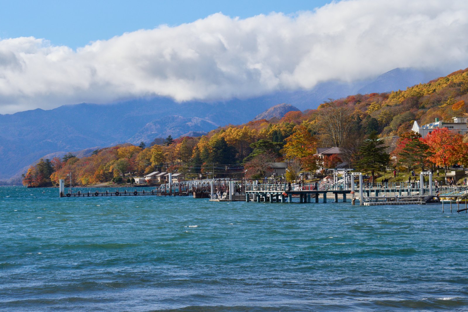 紅葉した山の斜面と湖畔の桟橋、手前に風で波立つ青緑色の中禅寺湖が写る