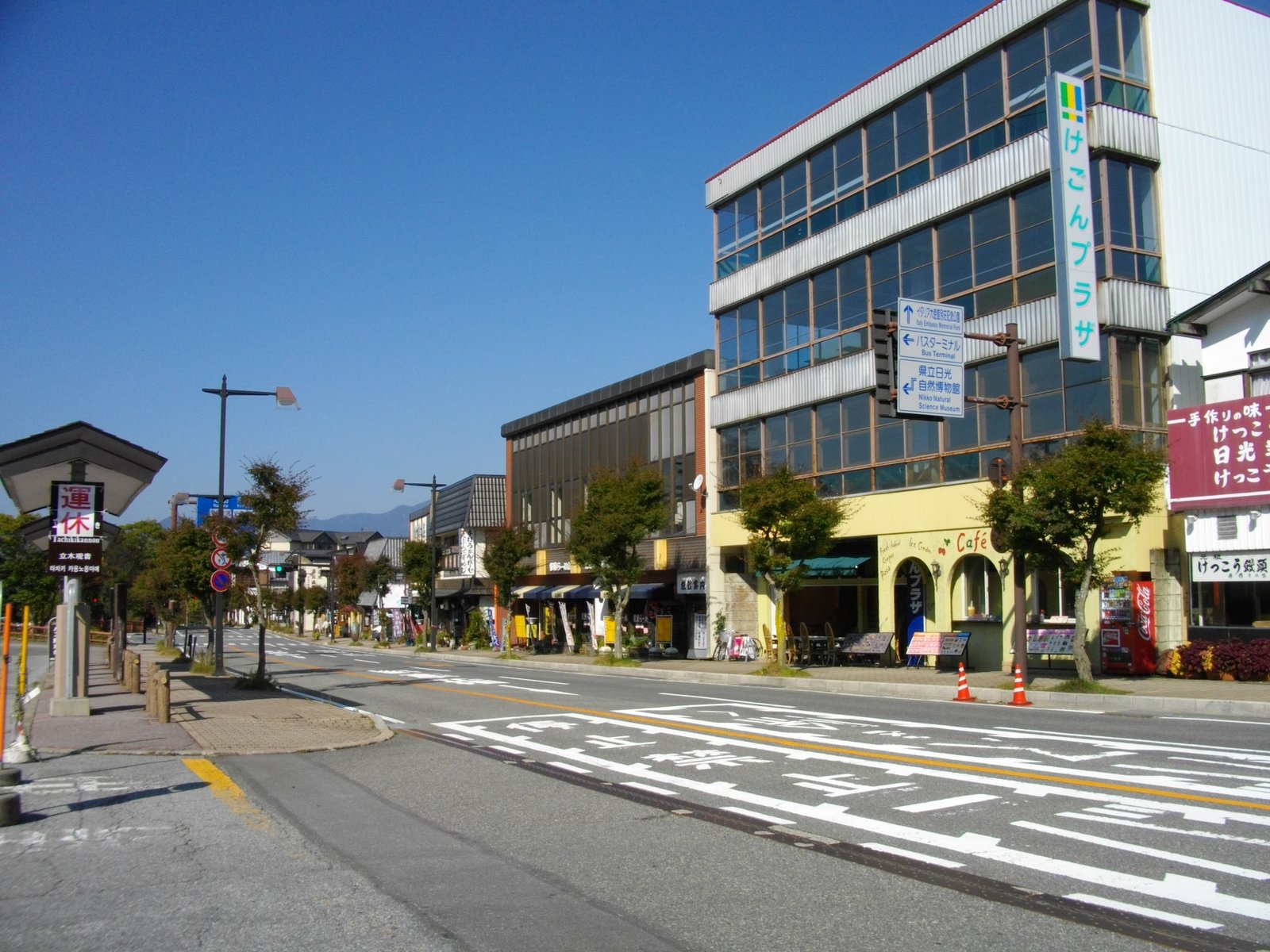 青空の下、片側にバス停の屋根と標識、反対側に「けごんプラザ」の看板を掲げた建物や店が並ぶ中禅寺温泉の通りと、奥に連なる山並み