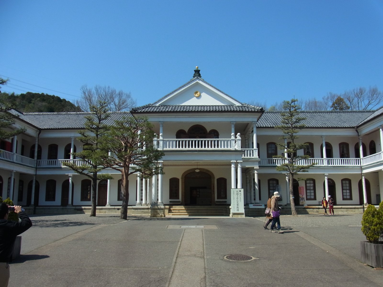 白い2階建ての洋風の建物。正面の中央に柱の並ぶバルコニーと三角の屋根があり、左右に長いベランダが延びる。前は広い広場