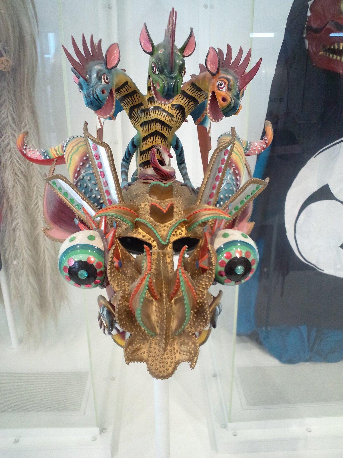 金色の顔に3つの獣の頭が乗った仮面。目玉を模した丸い飾りが左右に付き、色とりどりの鏡や布で装飾されている