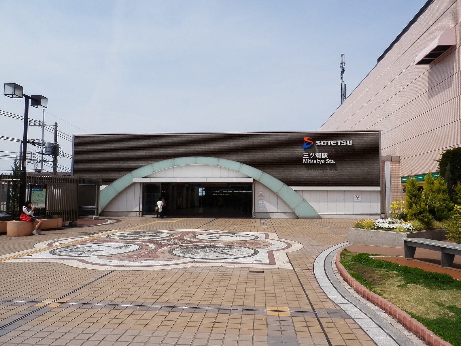 相鉄の三ツ境駅の入口。茶色の壁に緑色のアーチがかかり、前はタイル敷きの広場で花壇とベンチがある