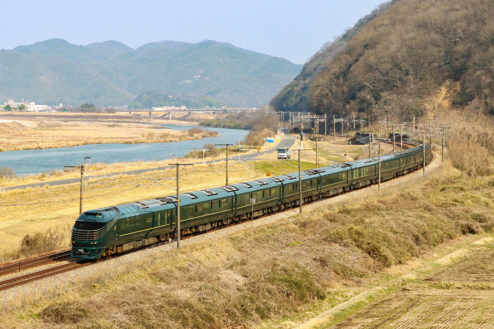 緑の車体に金色のラインが入った「TWILIGHT EXPRESS 瑞風」10両編成が、川沿いの築堤を走る