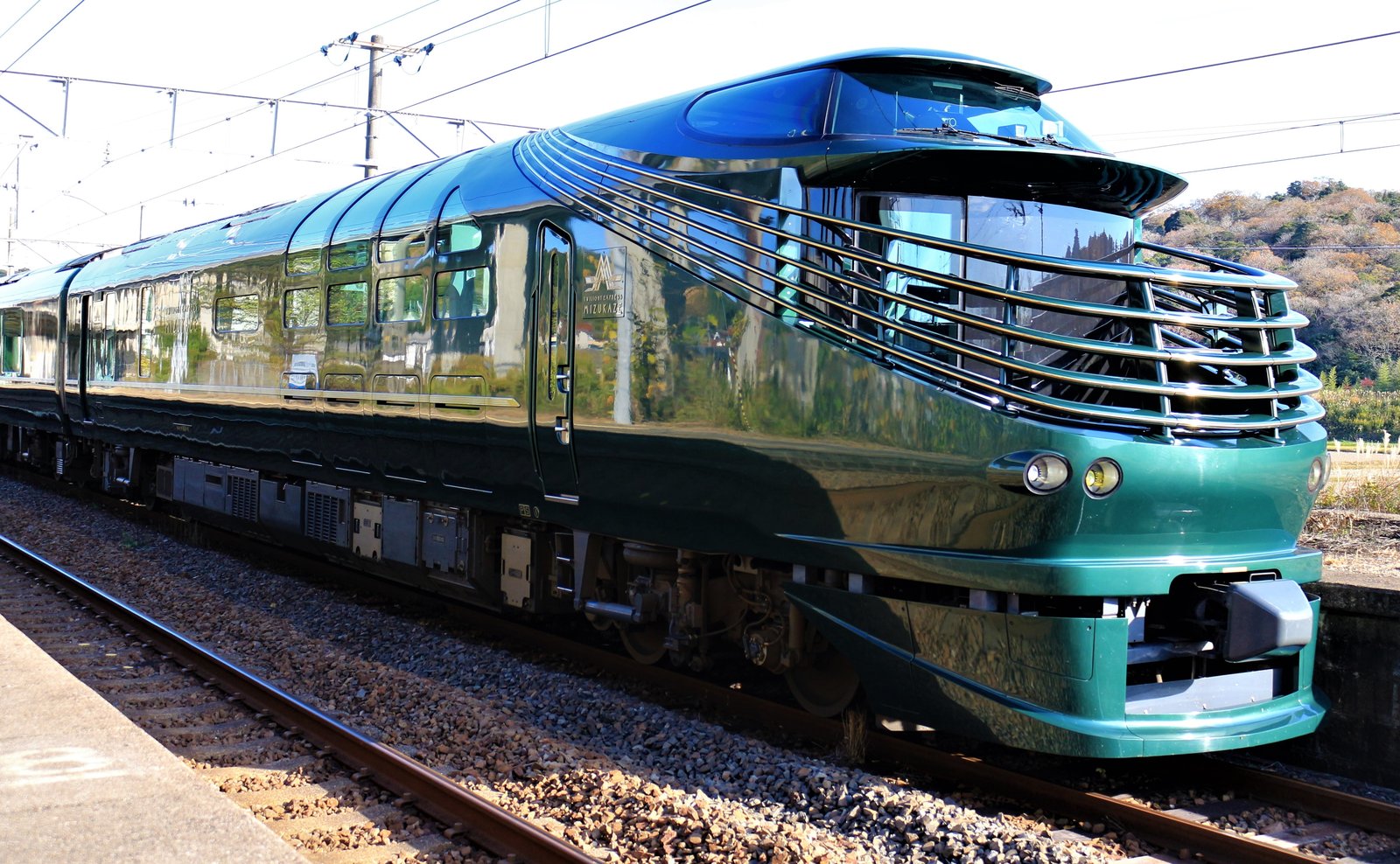 深い緑色の瑞風の先頭車。丸みのある先頭に金色の横線が何本も入り、側面に大きな窓が並ぶ。山陽本線の駅に停車中