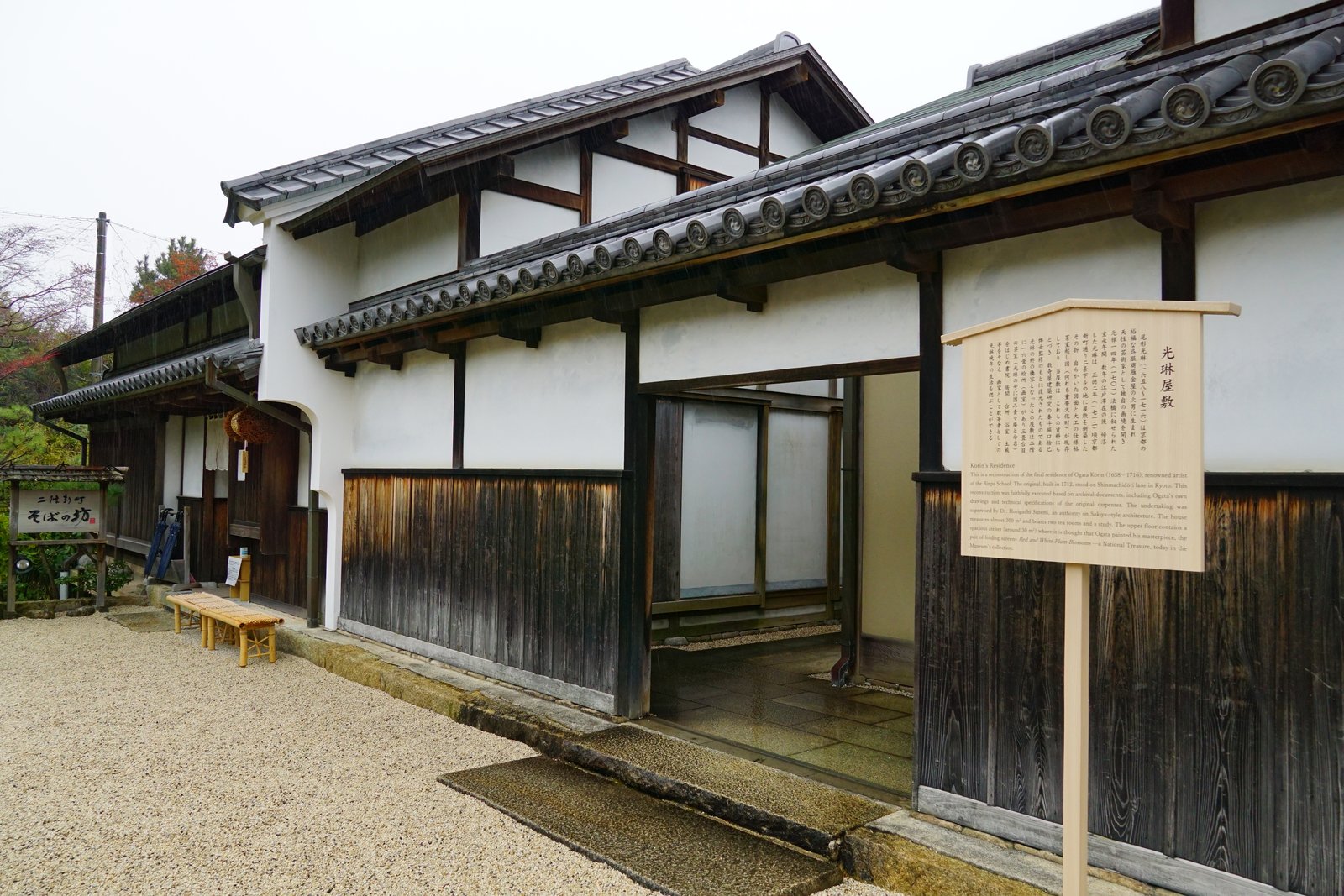 白壁と黒ずんだ板壁の2階建ての町家。瓦屋根の軒の前に「光琳屋敷」と書かれた木の説明板が立ち、左奥に「二條新町 そばの坊」の看板と竹の縁台が見える