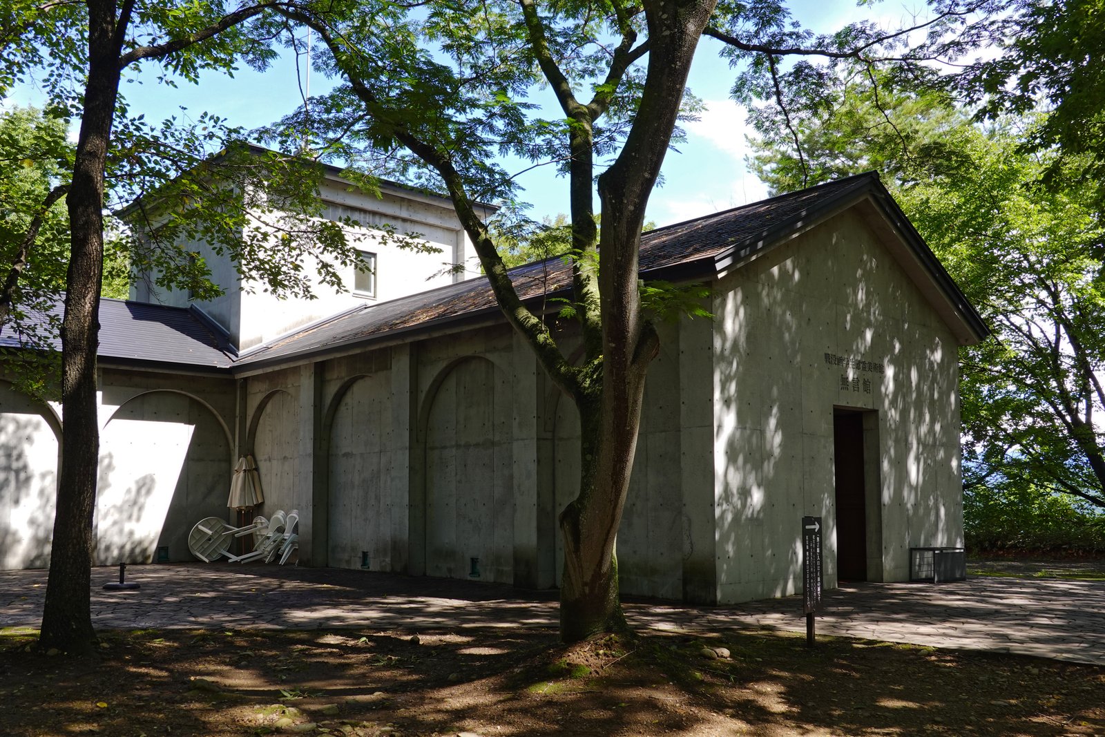 木立の間に建つ打ち放しコンクリートの無言館。切妻の壁に館名が刻まれ、右に四角い入口、左の壁には連続するアーチの跡がある
