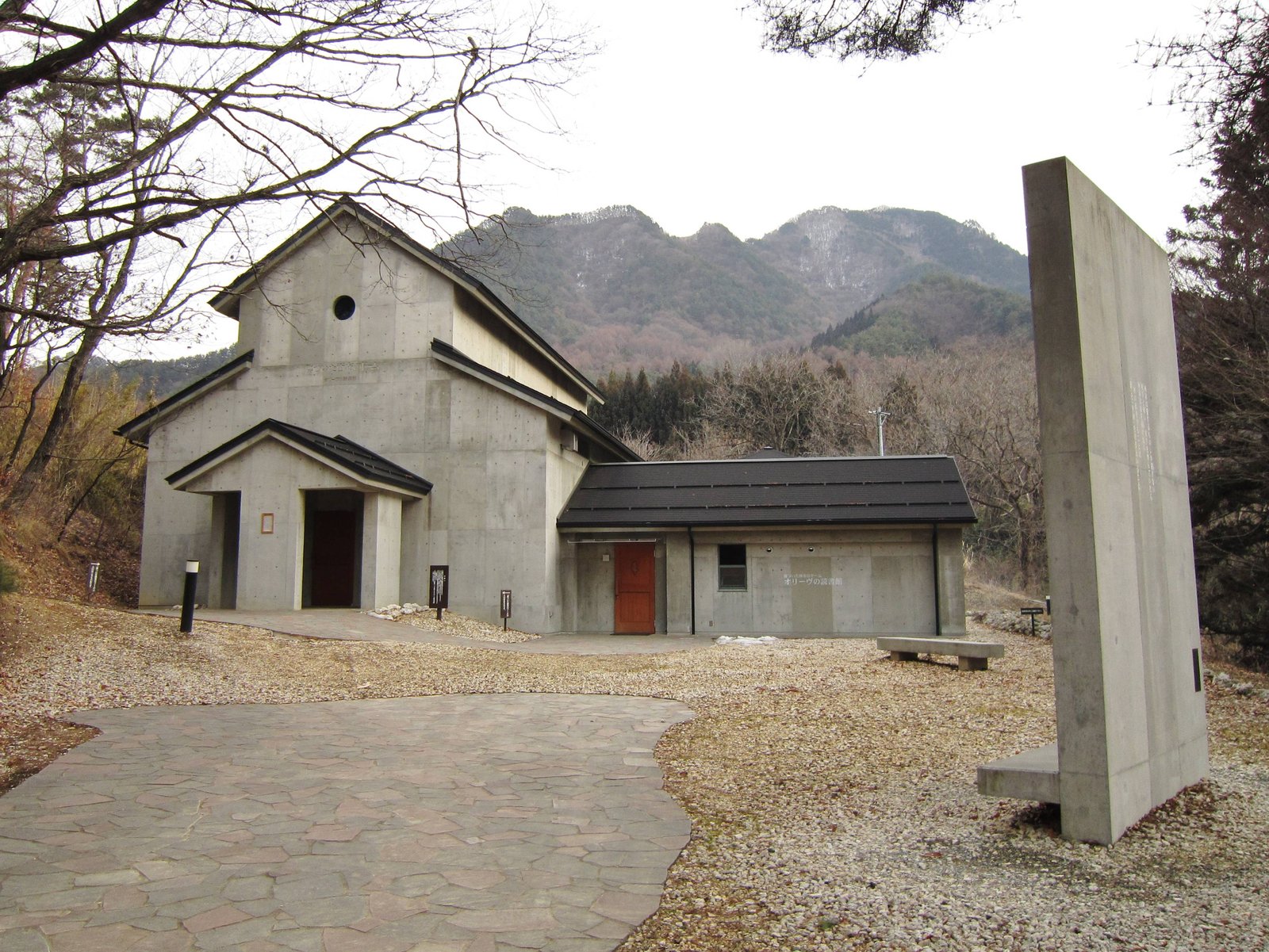 葉を落とした木々の向こうに山を背負って建つ、灰色のコンクリートの建物。右の低い棟には赤茶色の扉と「オリーヴの読書館」の文字があり、右端に大きなコンクリートの壁が立つ
