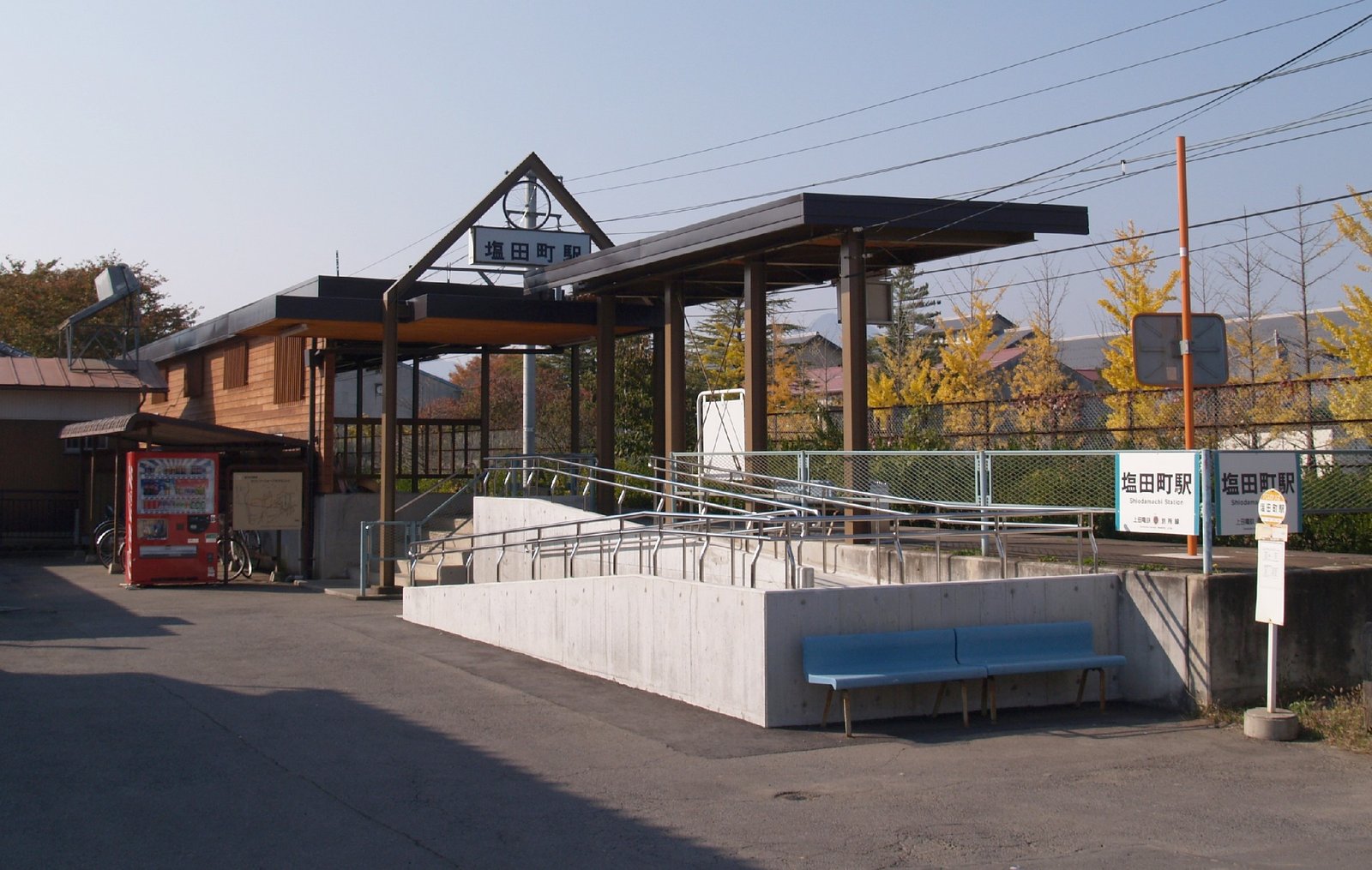木組みの屋根と三角形の看板を掲げた塩田町駅の駅舎。手前にスロープと青いベンチ、右に「塩田町駅」と書かれた駅名標が立つ