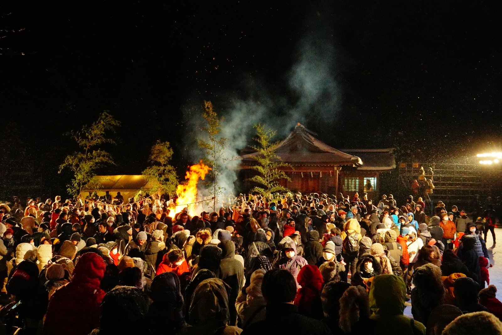 夜の真山神社の境内。社殿の前でかがり火が燃えて火の粉と煙が上がり、防寒着の観客が背を向けて取り囲んでいる