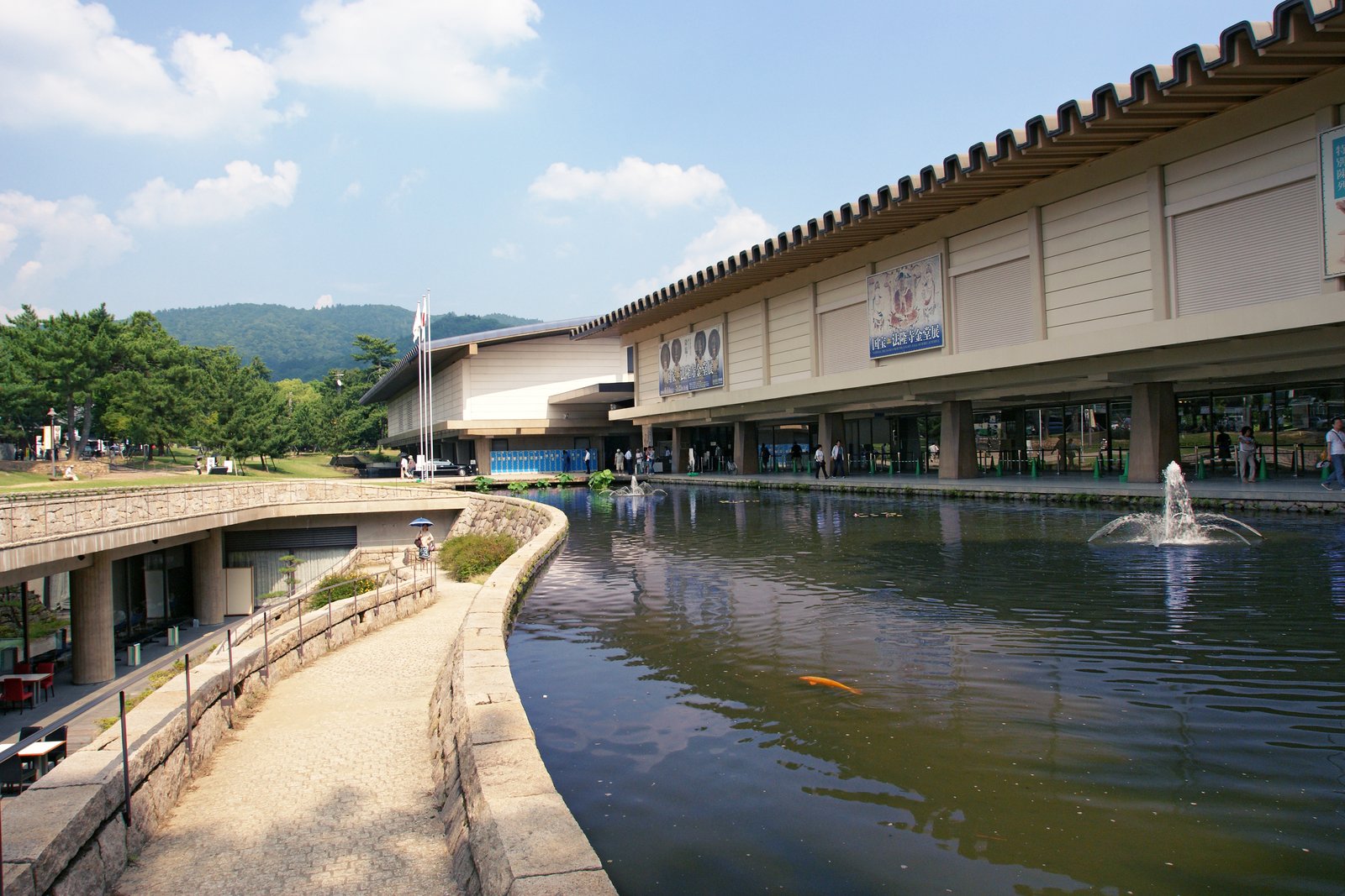 池に面して長く伸びる奈良国立博物館の新館。瓦屋根の低い建物の柱の下を人が歩き、壁に展覧会の垂れ幕が下がる。池では噴水が上がる