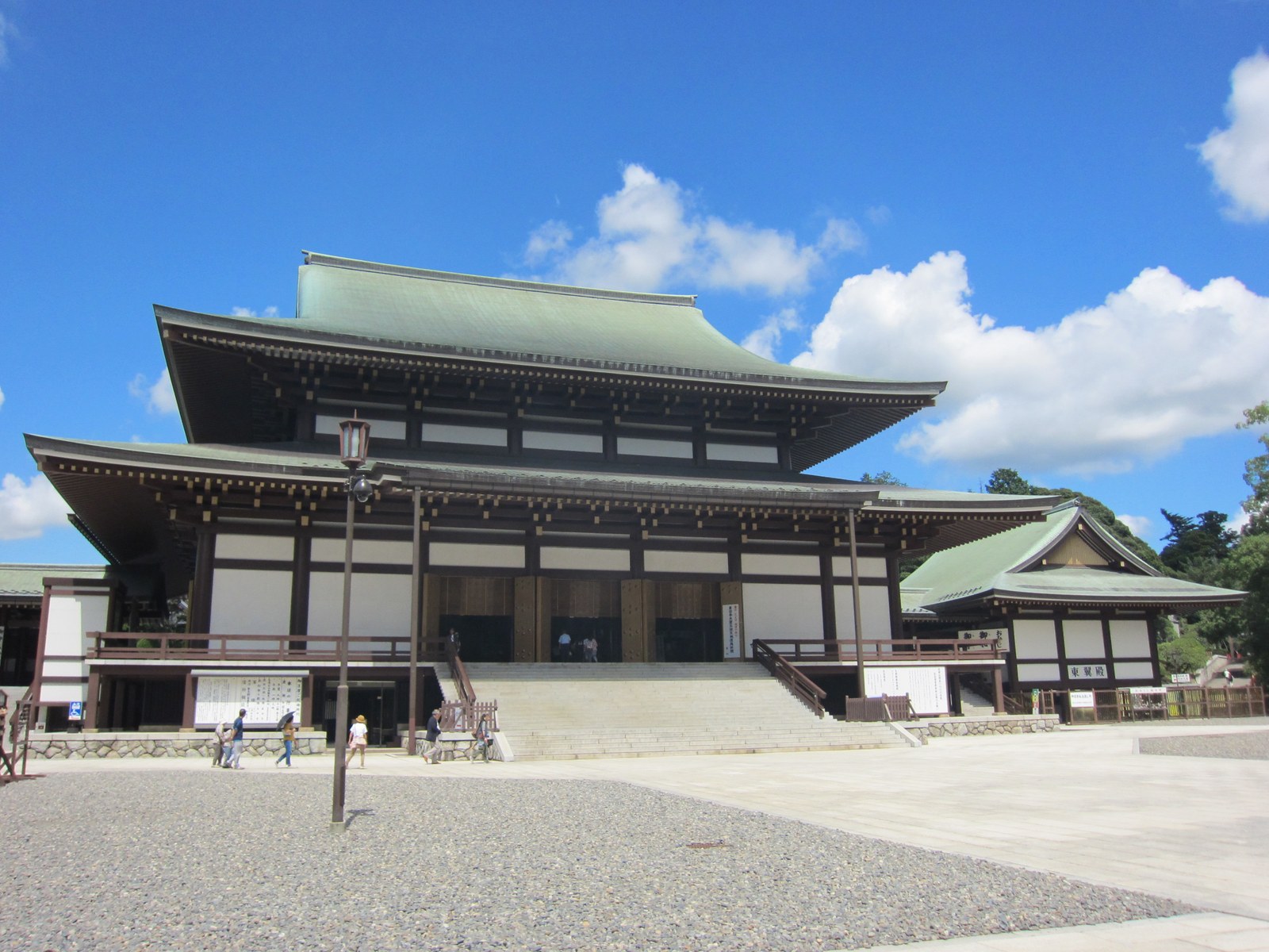 青空の下の成田山新勝寺の大本堂。二重の大きな屋根の堂の前に、広い石畳と砂利の庭が広がっている