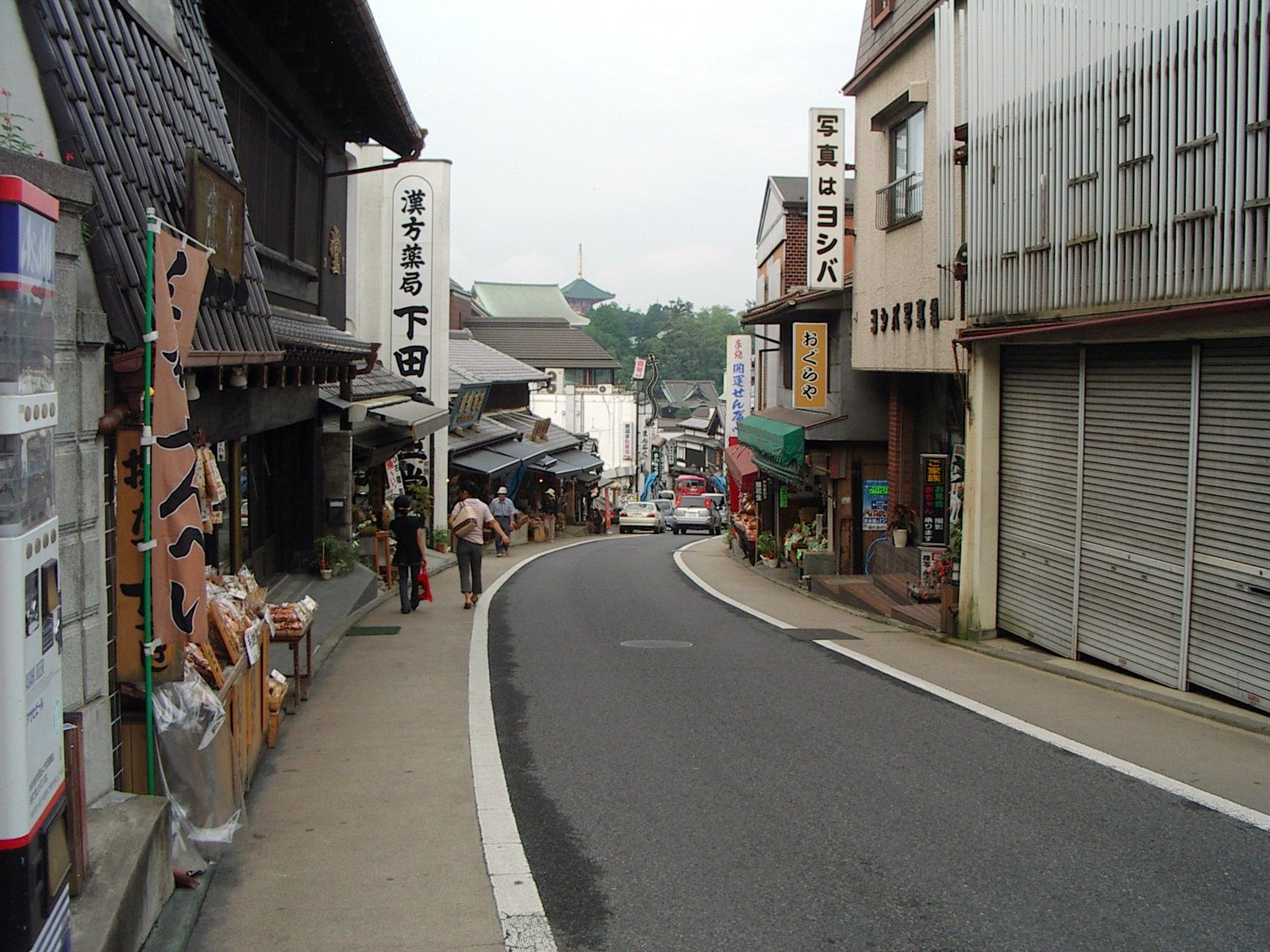 成田山の表参道。カーブした坂道の両側に土産物屋や薬局の看板が並び、奥に新勝寺の緑の屋根と塔の先が見える