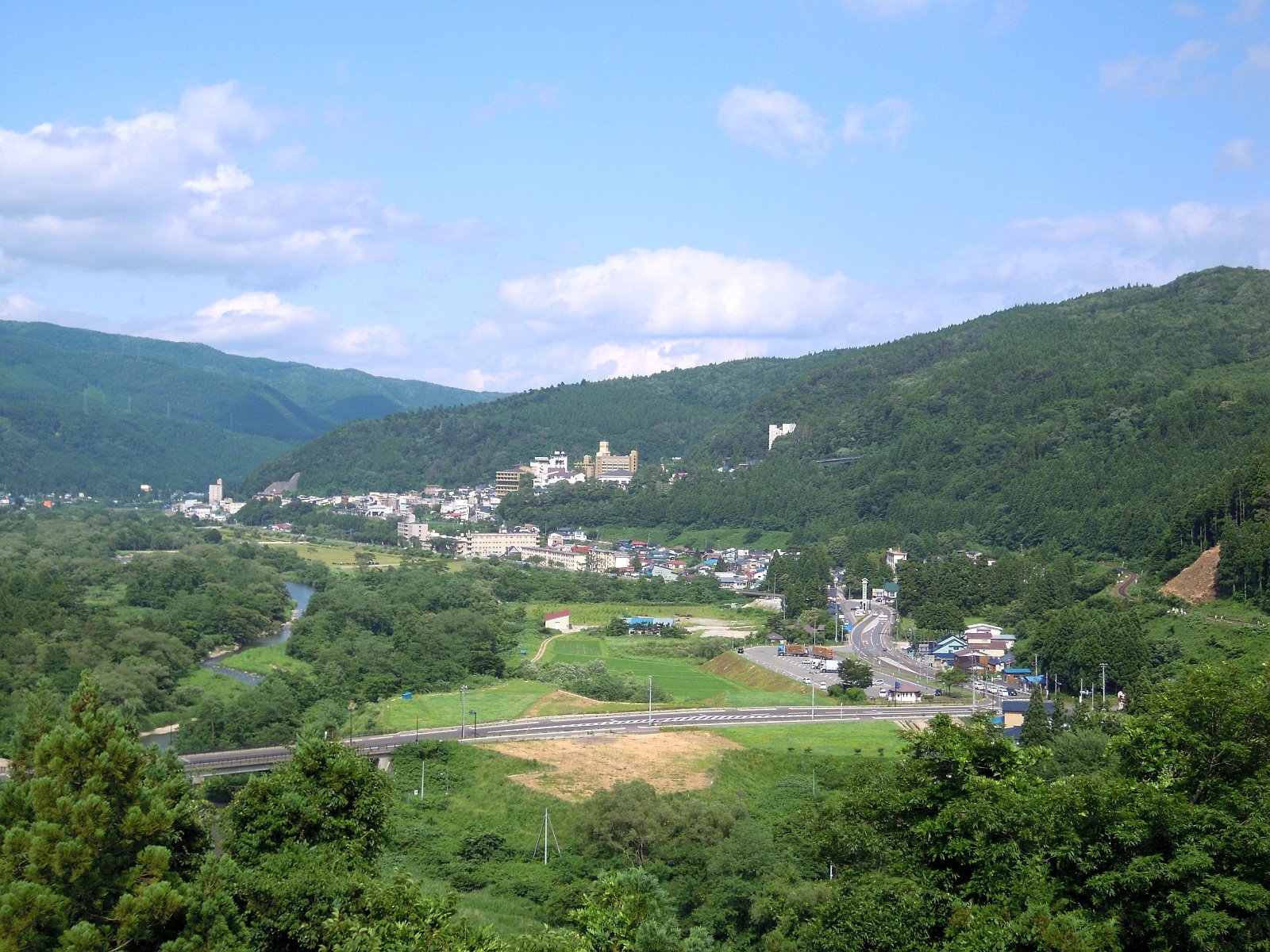緑の山に囲まれた谷あいに、白い旅館や住宅が集まる鳴子温泉の町並み。手前には田んぼと道路、左に川が見える。