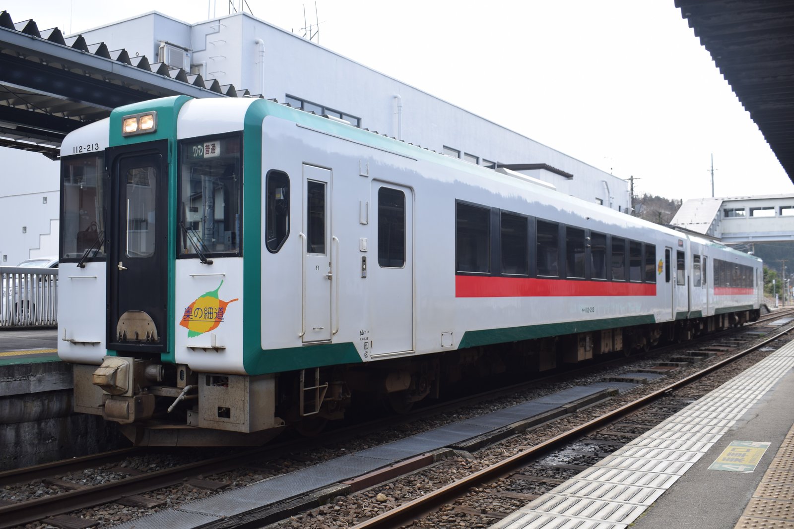 鳴子温泉駅のホームに停まる陸羽東線のキハ112形。白い車体に緑と赤の帯があり、運転席の脇に「奥の細道」のロゴが付いている