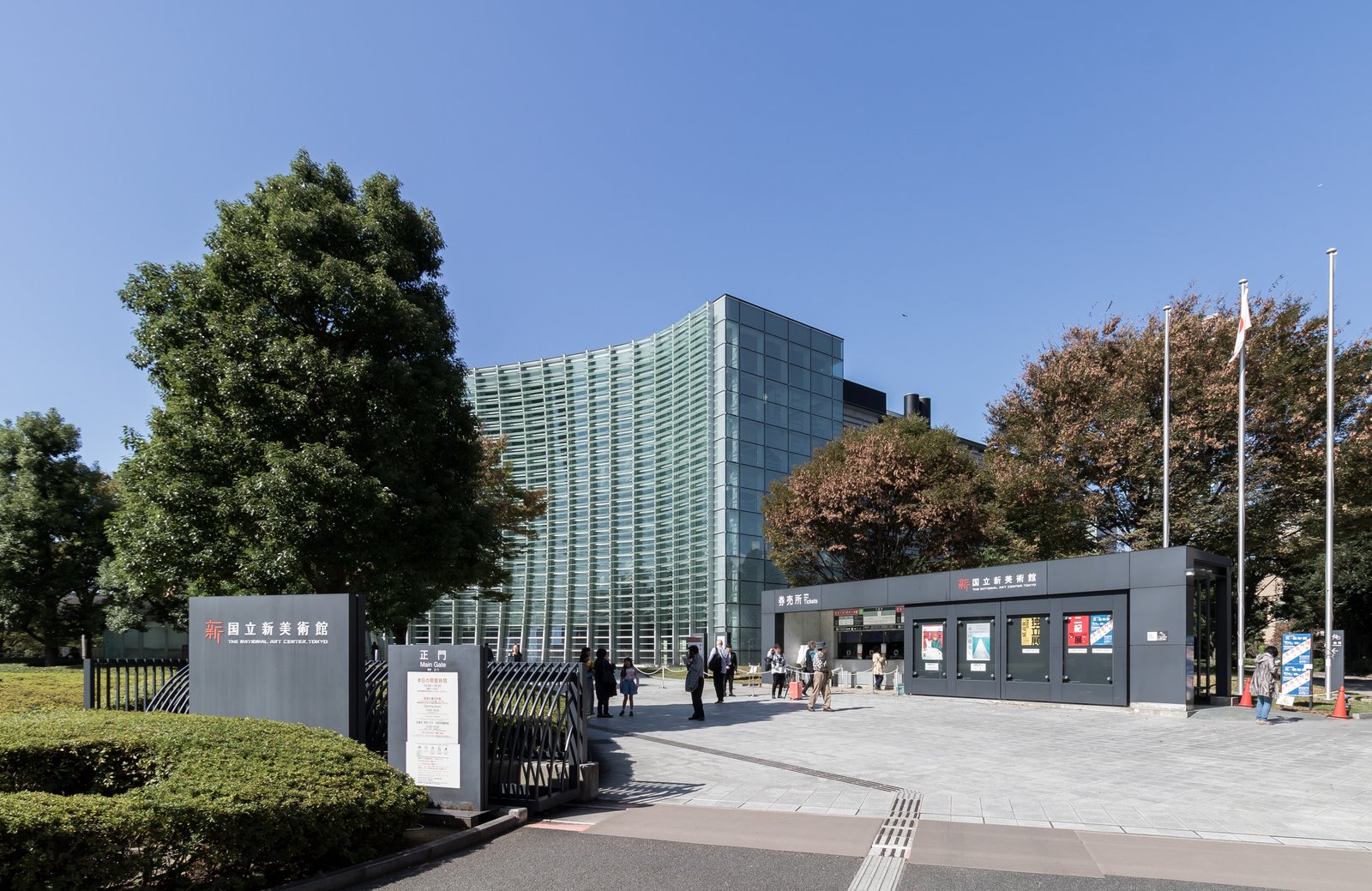 国立新美術館の正門と券売所の向こうに、波のように湾曲した緑がかったガラスのカーテンウォールが立つ。手前と両脇に木が茂り、空は晴れている