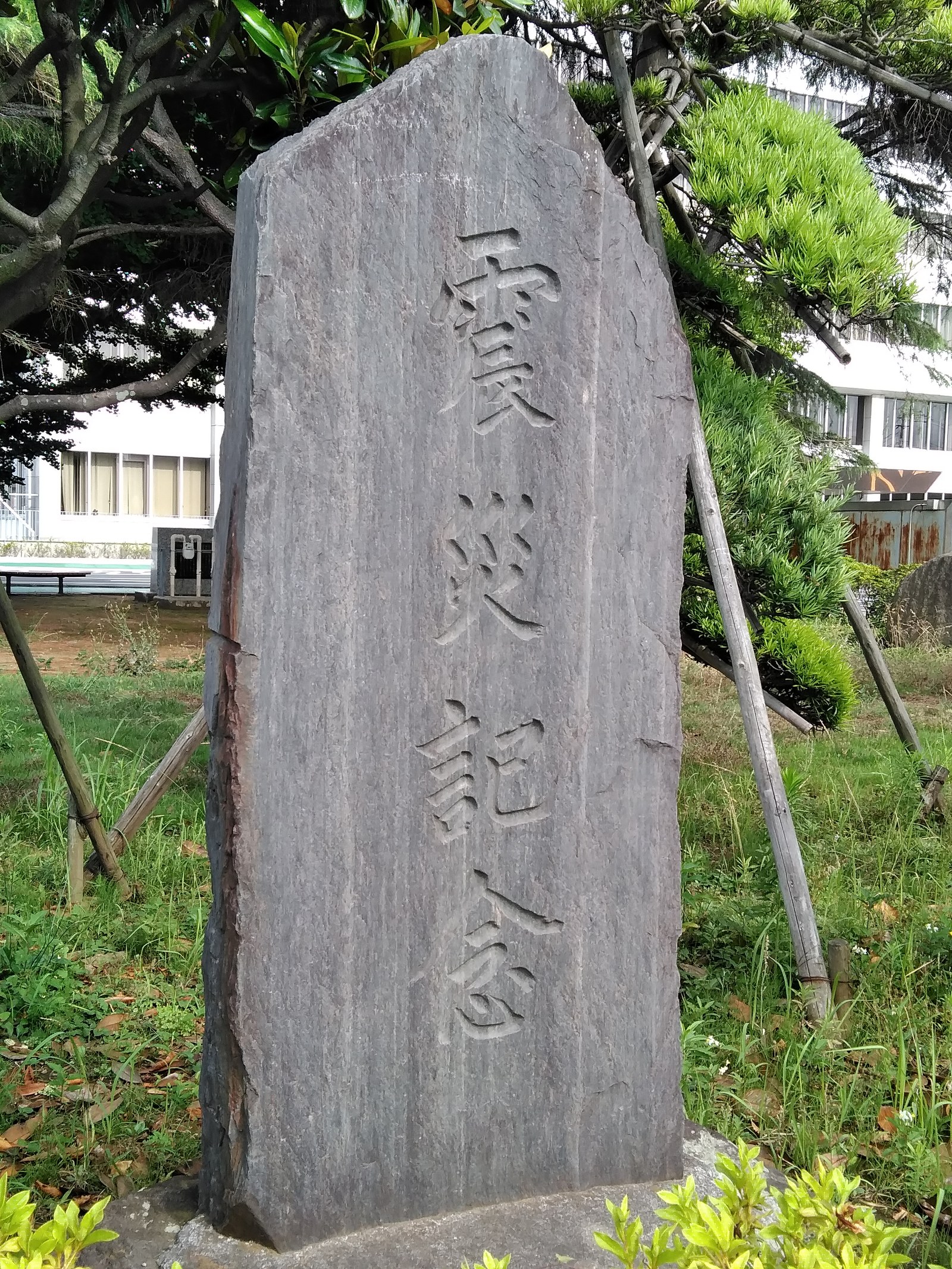 自然石を縦に立てた石碑。表面に「震災記念」の文字が縦書きで彫られ、周囲は芝生と庭木に囲まれている（千葉県鎌ケ谷市）