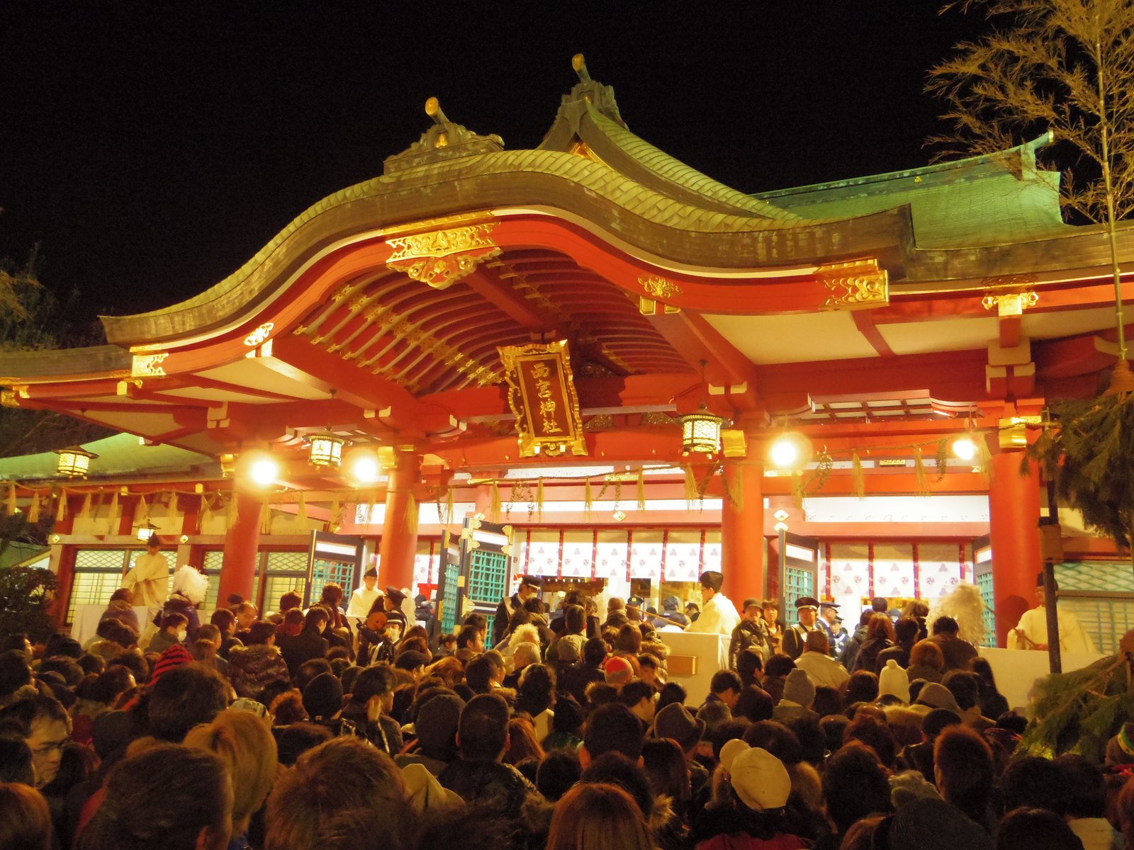 夜、明かりに照らされた朱塗りの拝殿。「西宮神社」の額の下に白い装束の神職が立ち、手前を大勢の参拝者が埋めている