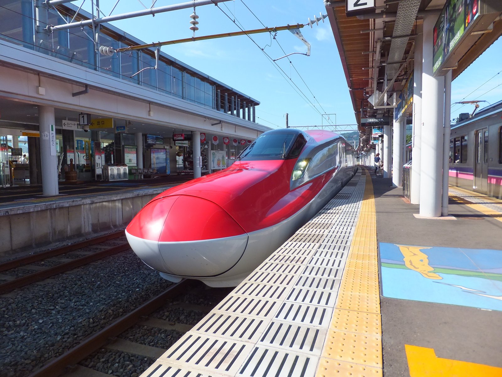 田沢湖駅のホームに停まる、赤い車体のE6系秋田新幹線こまち。左に駅舎、右に別の路線の電車が見える
