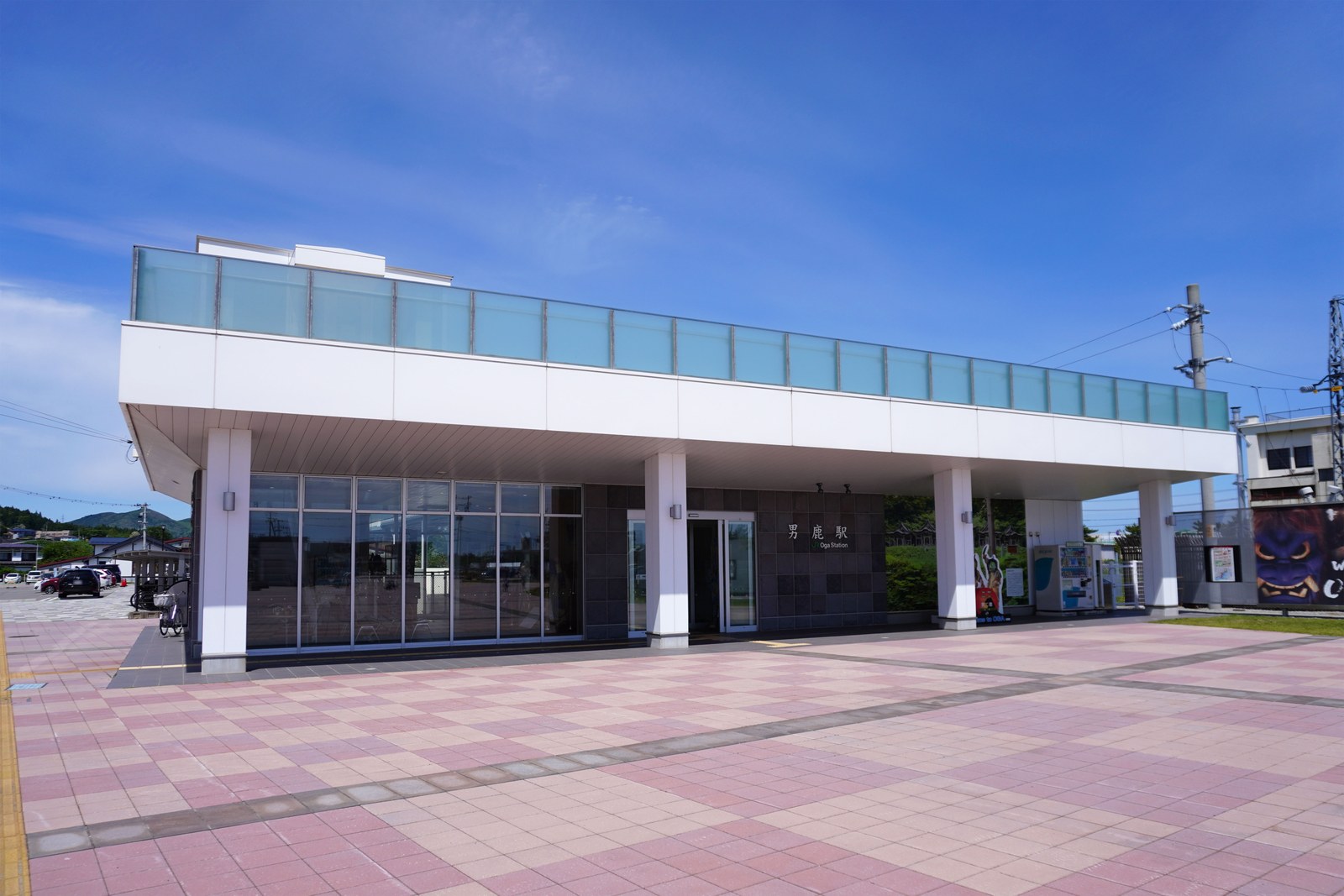 JR 男鹿駅の駅舎。平たい白い屋根の上に展望デッキがあり、前に広場が広がっている