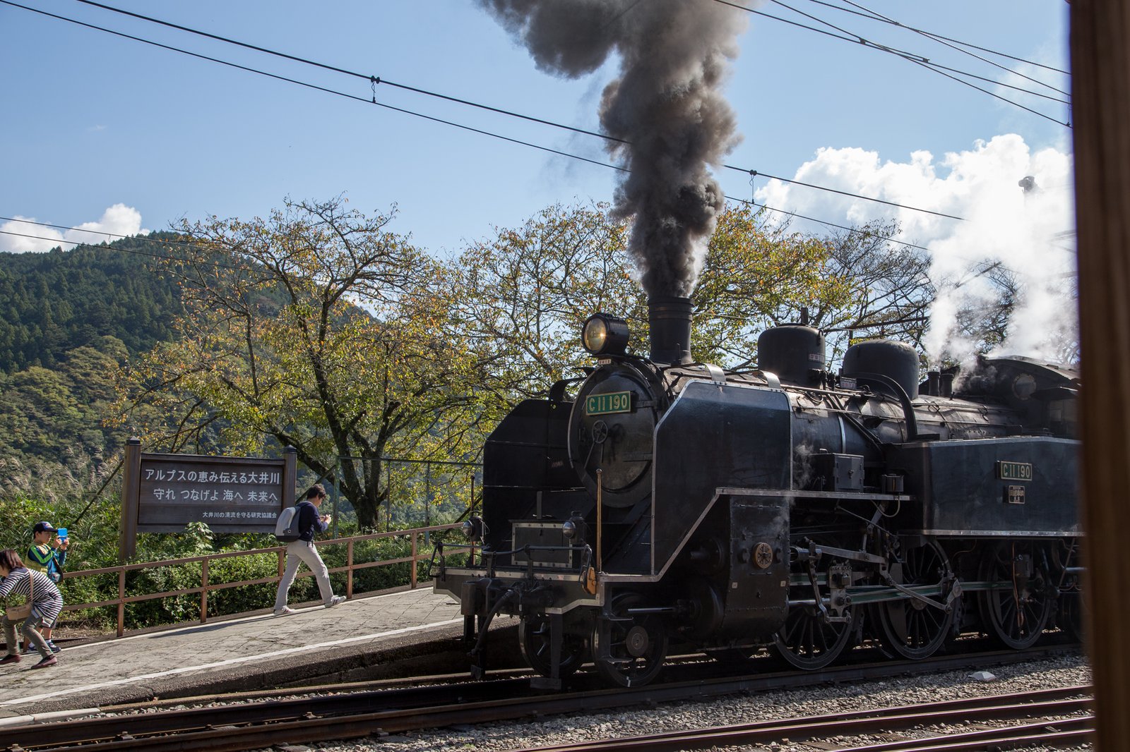 家山駅のホームに停まる大井川鐵道の黒い蒸気機関車 C11 190。煙突から黒い煙が上がり、後ろに山と「アルプスの恵み伝える大井川」の看板