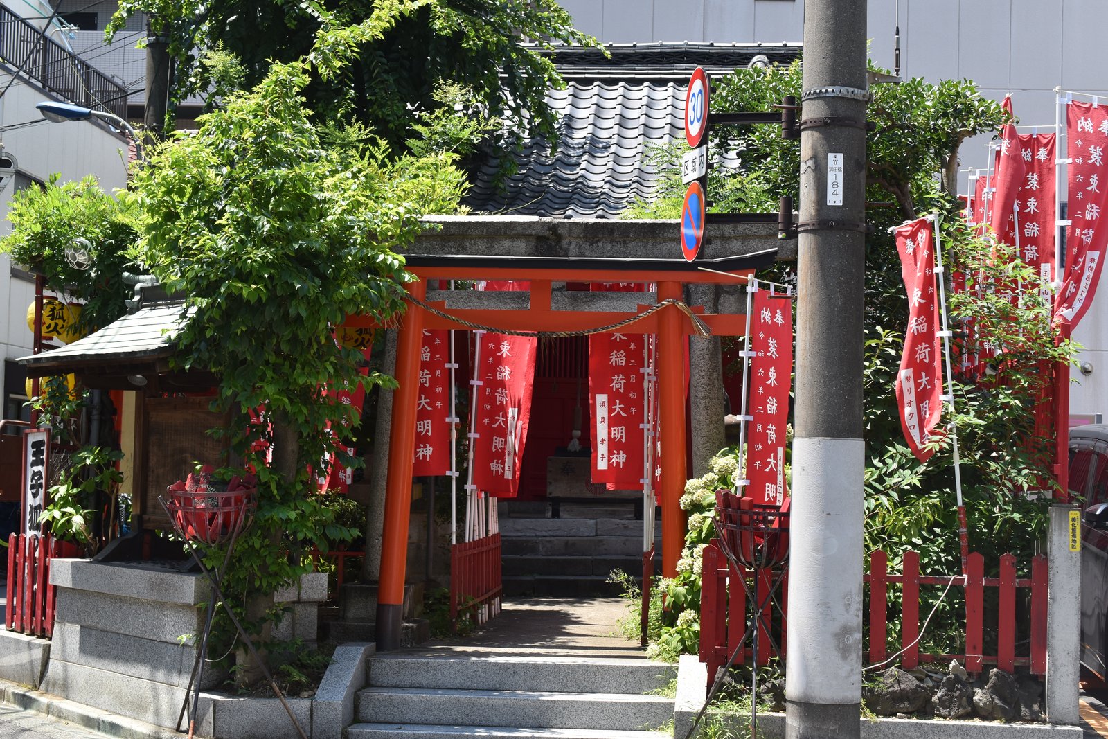 住宅街の角に建つ装束稲荷神社。朱の鳥居の奥に小さな社殿があり、「装束稲荷大明神」と書かれた赤いのぼりが並ぶ