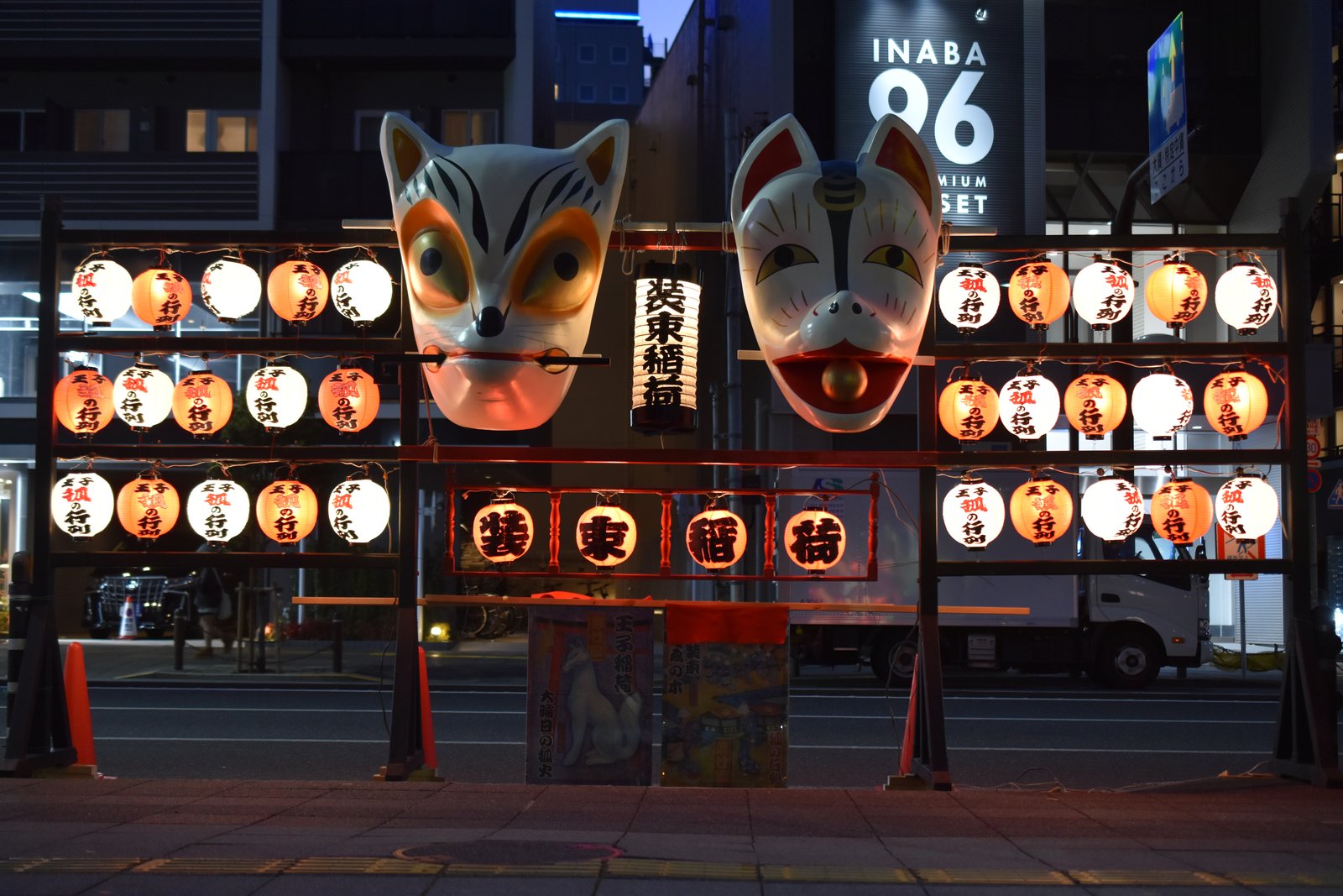 夜の街路に据えられた櫓。白い大きな狐の面が2つ並び、「王子狐の行列」と書いた赤と白の提灯と「装束稲荷」の看板が掛かる