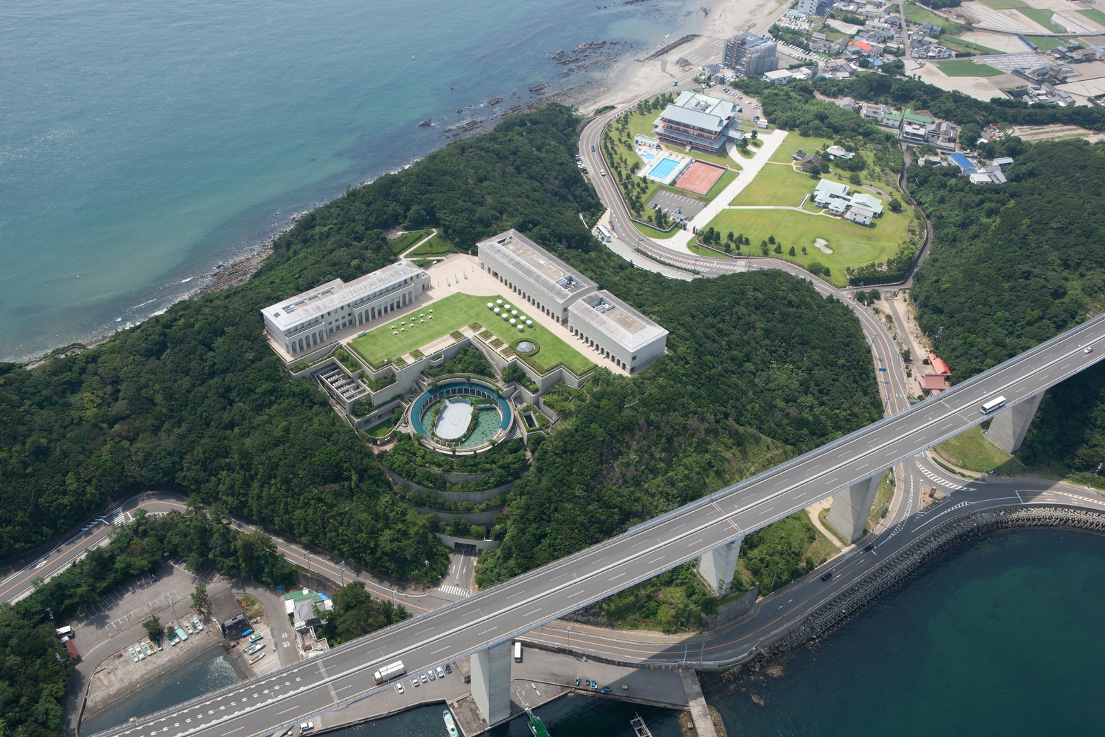上空から見た大塚国際美術館。海に面した緑の山の上に、四角い建物と芝生の広場、円形の池がある。手前を道路の高架が横切る