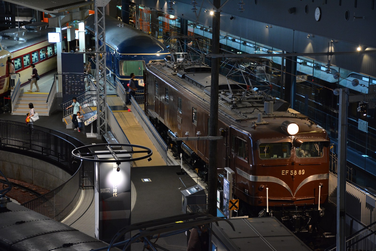 鉄道博物館 車両ステーション1Fの展示。手前に茶色のEF58形電気機関車89号機、奥に青い寝台特急の客車と黄色帯の電車、見学用の通路と階段