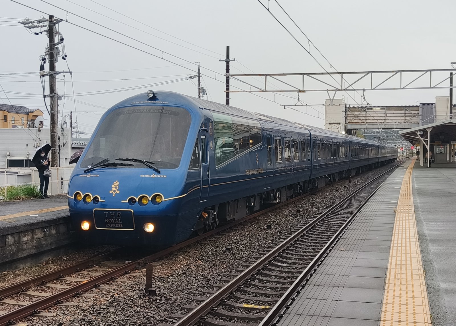 雨の東海道本線 新居町駅のホームに停まるTHE ROYAL EXPRESSの先頭車。ロイヤルブルーの車体に王冠の紋章と金色の文字、前面に丸いライトが6つ並ぶ