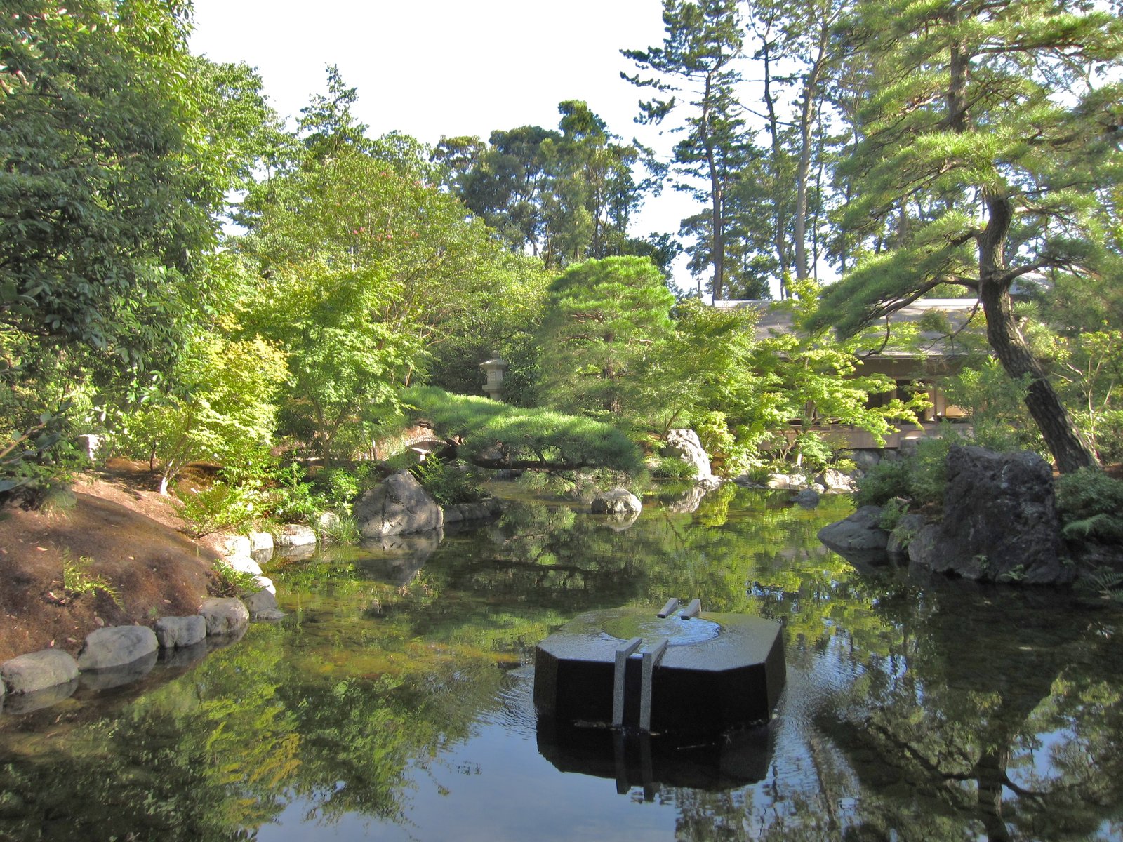 神嶽山神苑の池。松や広葉樹に囲まれた池に岩が配され、水面に木々が映り、奥に木造の建物の屋根が見える