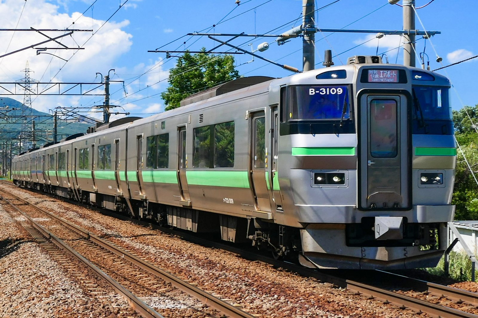 銀色の車体に緑の帯の電車が線路を走ってくる。先頭の行き先表示は「特別快速 エアポート」