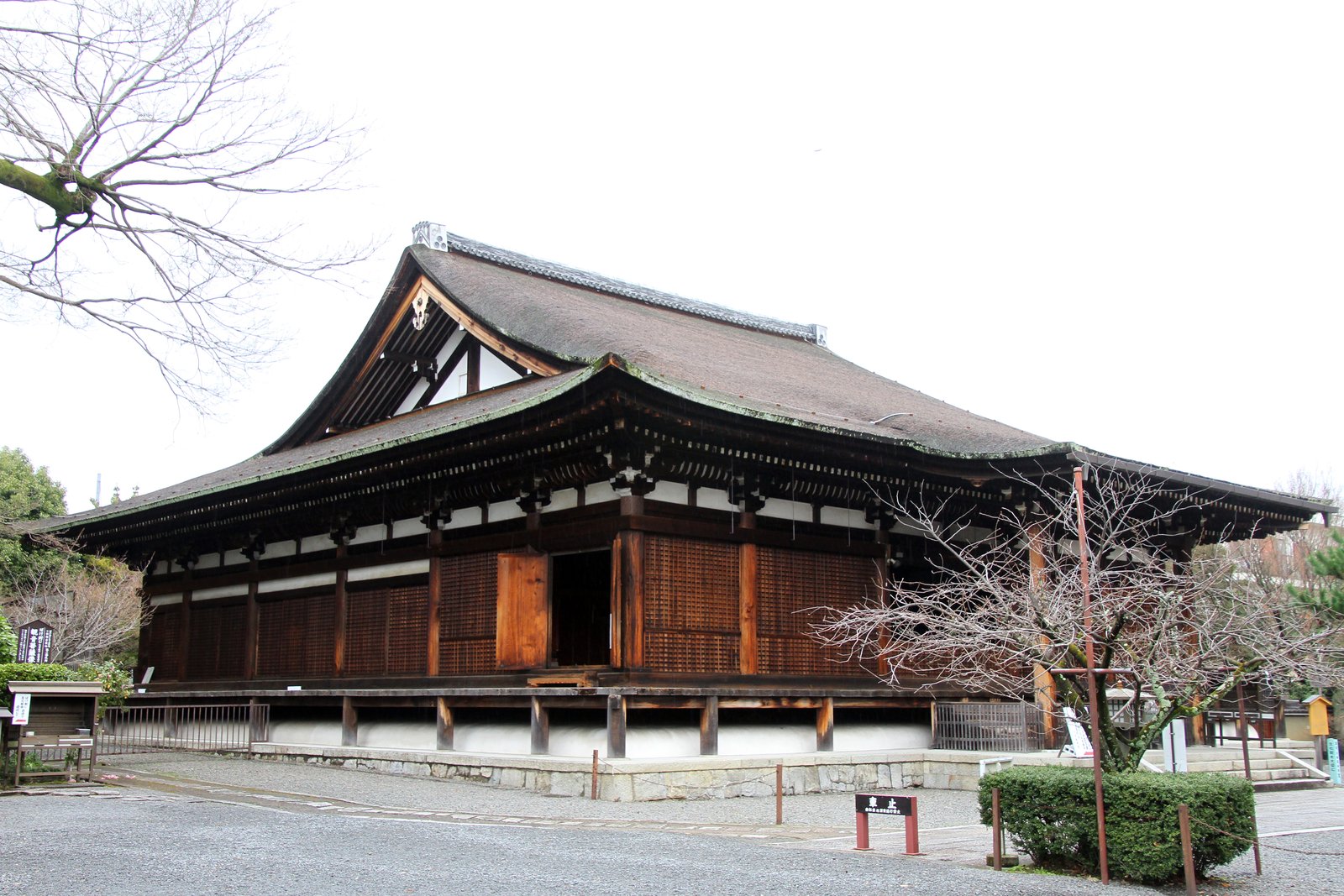 冬枯れの木に囲まれた千本釈迦堂（大報恩寺）の本堂を斜め前から見た写真。檜皮葺の入母屋の大屋根の下に格子の戸が並び、板戸が1枚開いている