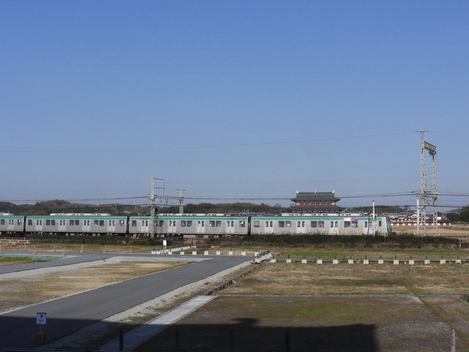 平城宮跡の草地の向こうを銀色に緑の帯の近鉄電車が横切り、その奥に朱色の柱と緑の瓦の第一次大極殿が見える