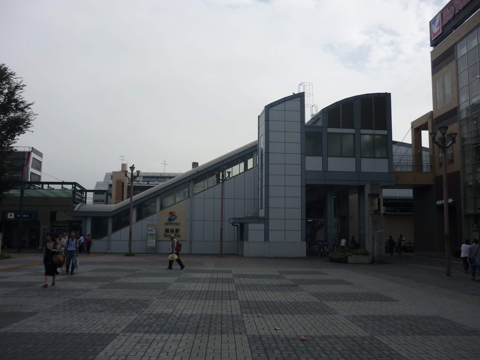 相鉄の瀬谷駅の北口。石畳の広い広場の奥に、斜めの屋根の通路とグレーの駅舎があり、右に商業ビルが建つ