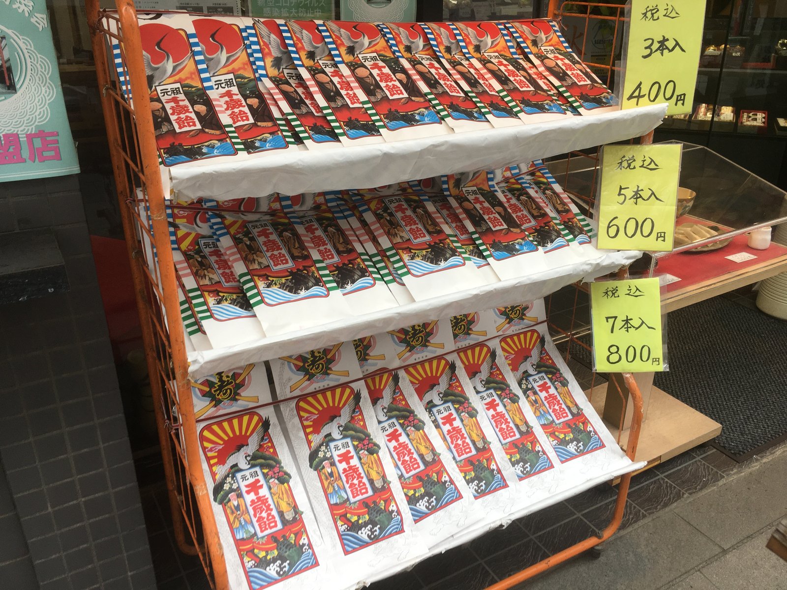 店先の橙色のラックに並ぶ千歳飴の袋。鶴と日の出、松を描いた赤と白の縦長の袋が、本数ごとに三段に分けて入れてある