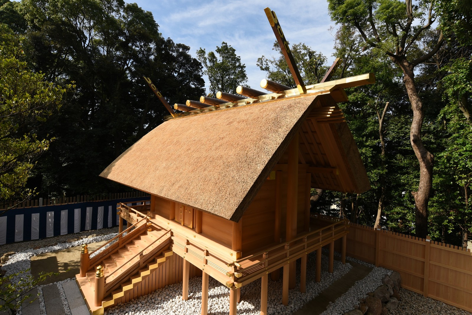 伊勢山皇大神宮の茅葺き屋根の本殿を斜め下から見た外観。木々に囲まれ、白い玉砂利の上に建つ