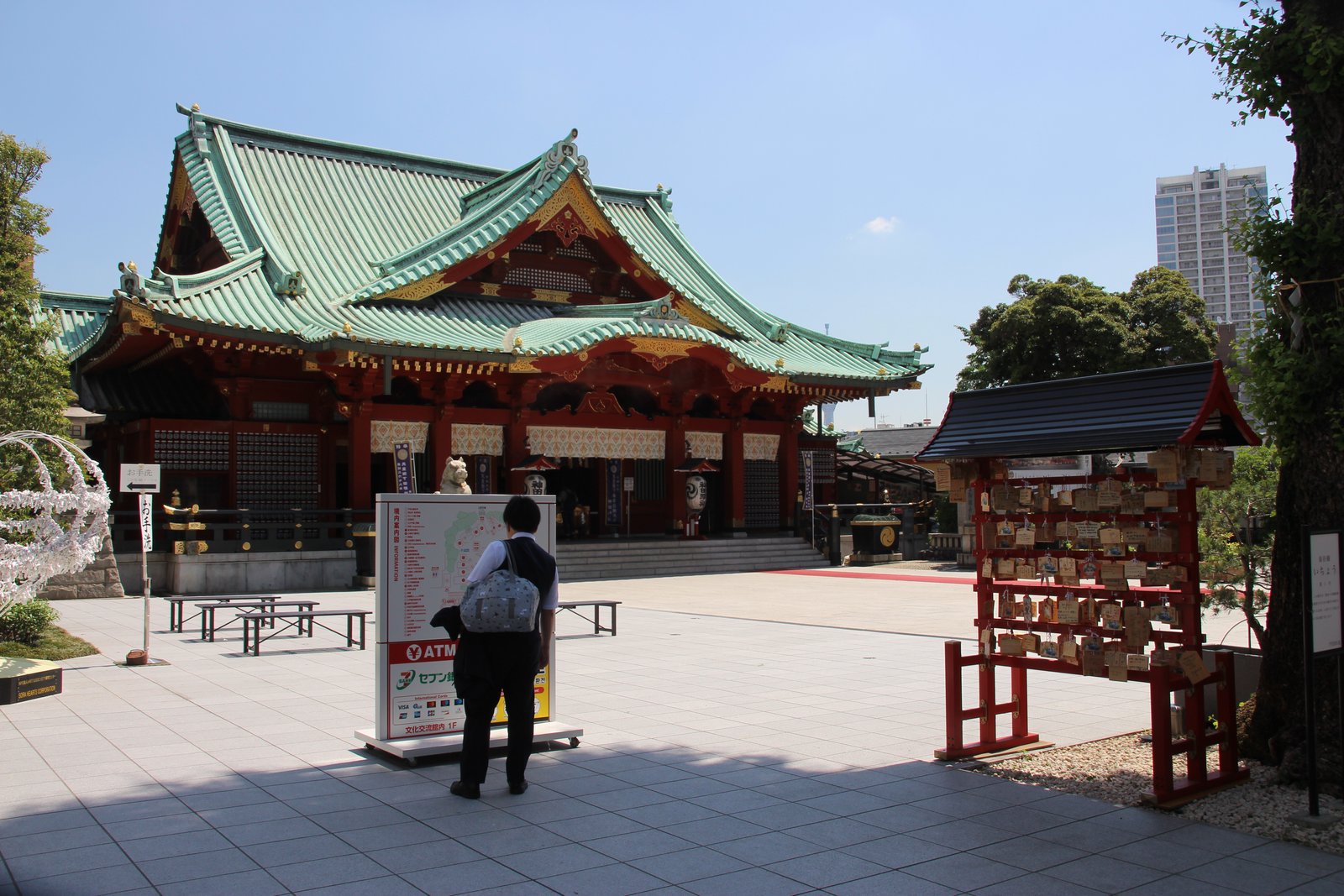 神田明神の社殿。緑の屋根に朱塗りの柱の社殿で、手前の広場に境内の案内板と絵馬掛け、奥に高層ビルが見える