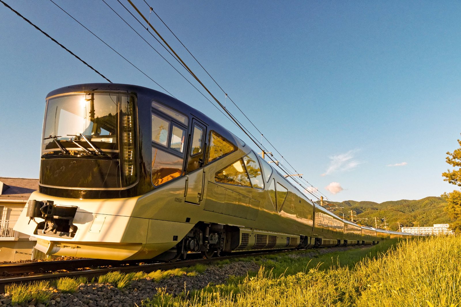 夕日を受けて走るTRAIN SUITE 四季島の先頭車。シャンパンゴールドの車体に、上へ大きく傾いたガラス張りの展望窓が並び、山並みの手前に10両が続く