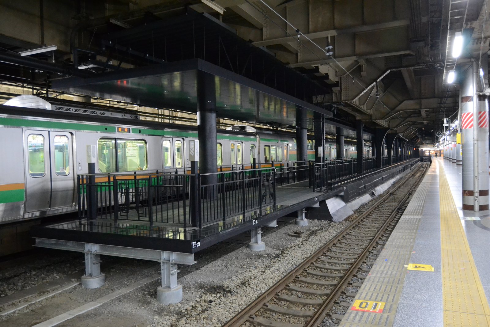 上野駅の13.5番線ホーム。線路の間に設けられた黒い低いホームで、黒い柵と屋根が付き、奥に緑とオレンジの帯の通勤電車が停まっている