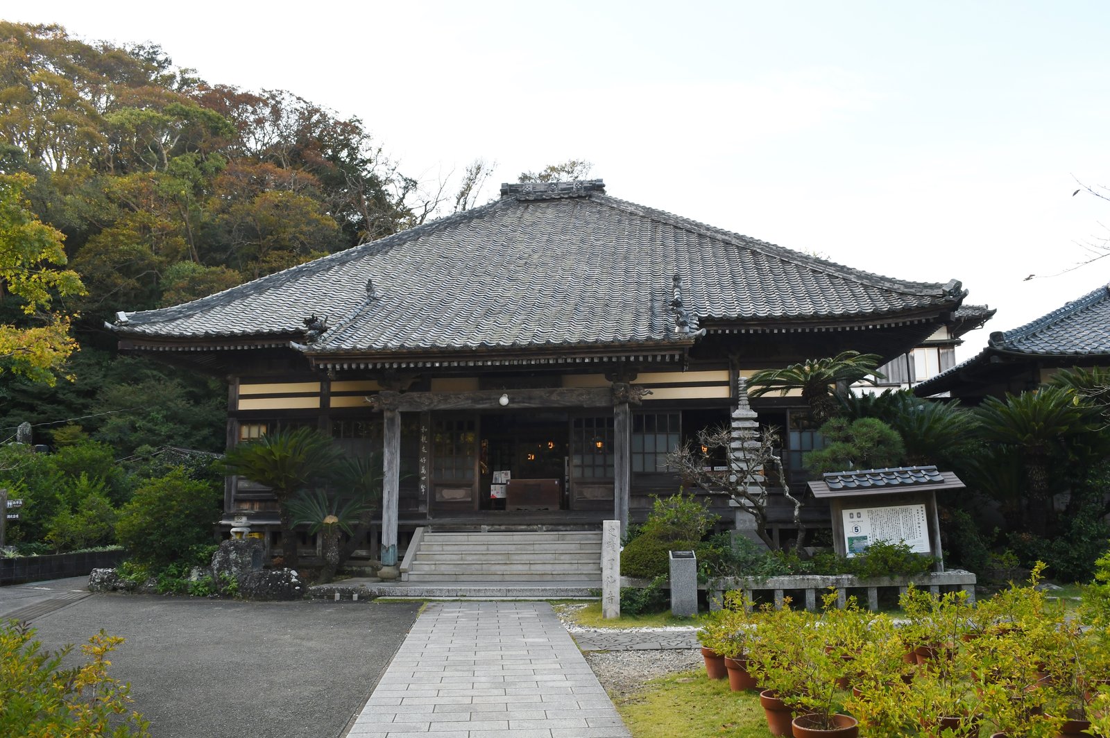 秋の木々を背にした了仙寺の本堂。入母屋の瓦屋根と石段、手前に石畳の参道と鉢植えが見える