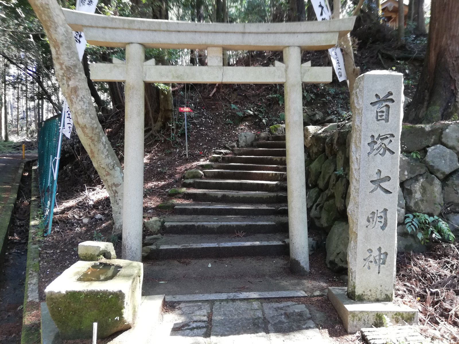 首塚大明神の入口。杉林の中に石の鳥居と石段があり、右に「首塚大明神」と彫られた石の碑、左に手水鉢が置かれている
