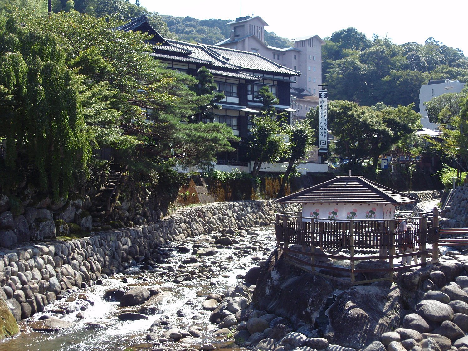 石積みの護岸の桂川の岩場に、花の絵が描かれた白壁と切妻屋根の独鈷の湯が建つ。対岸に黒い瓦屋根の旅館と緑の木々が見える。